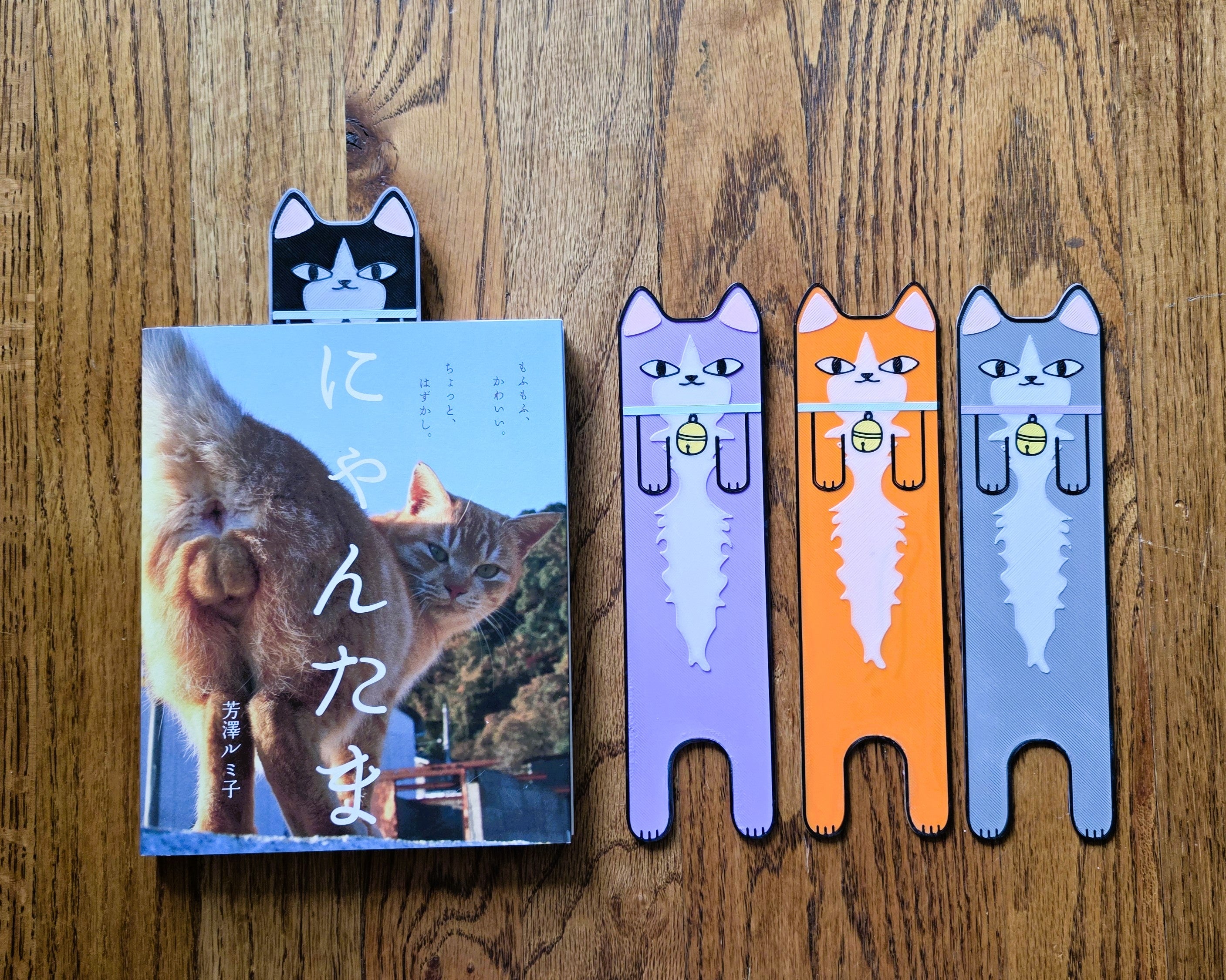 Cat-shaped bookmarks in various colors next to a book on a wooden surface