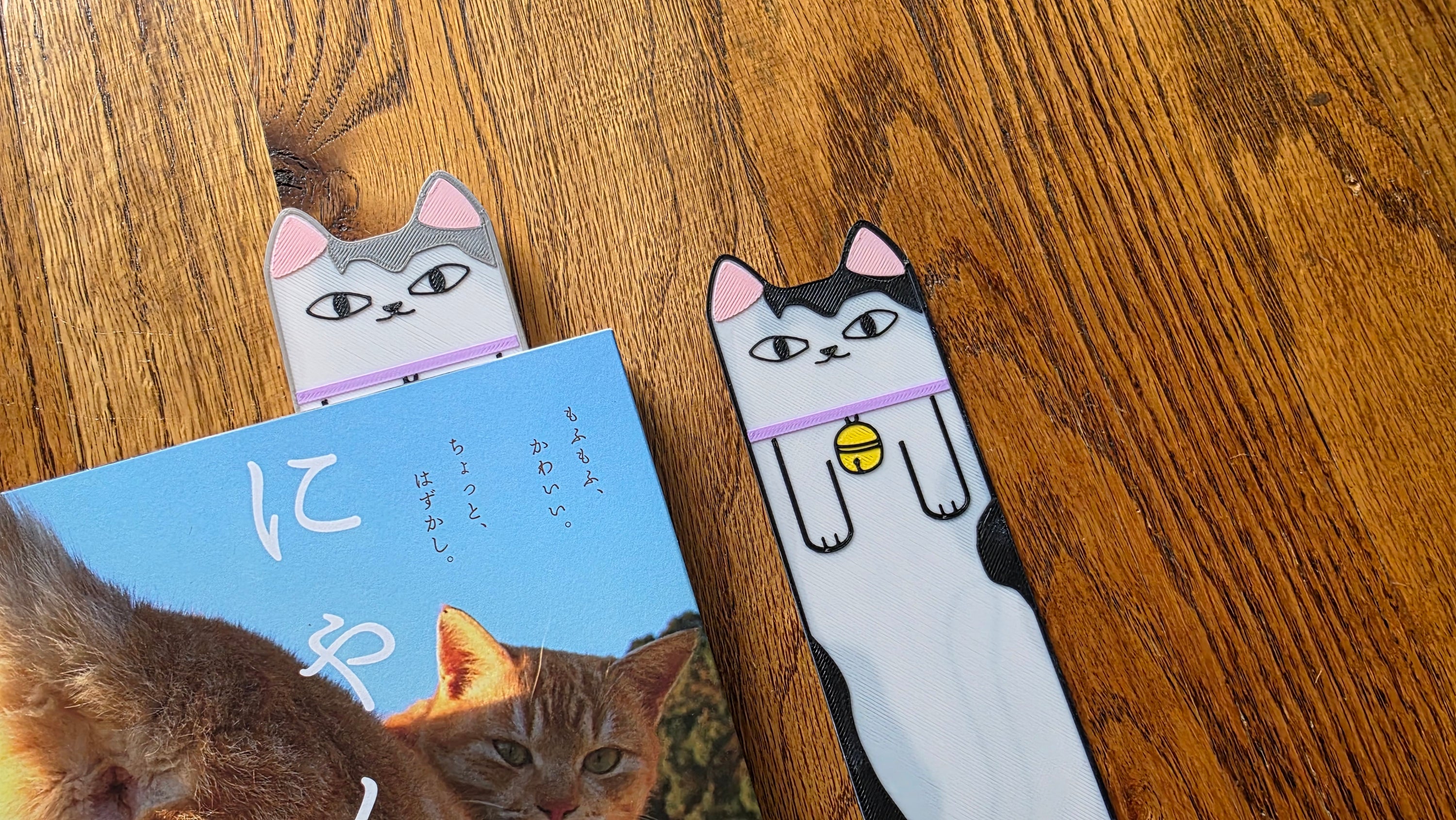 Two cat-shaped bookmarks on a wooden surface with a book underneath.