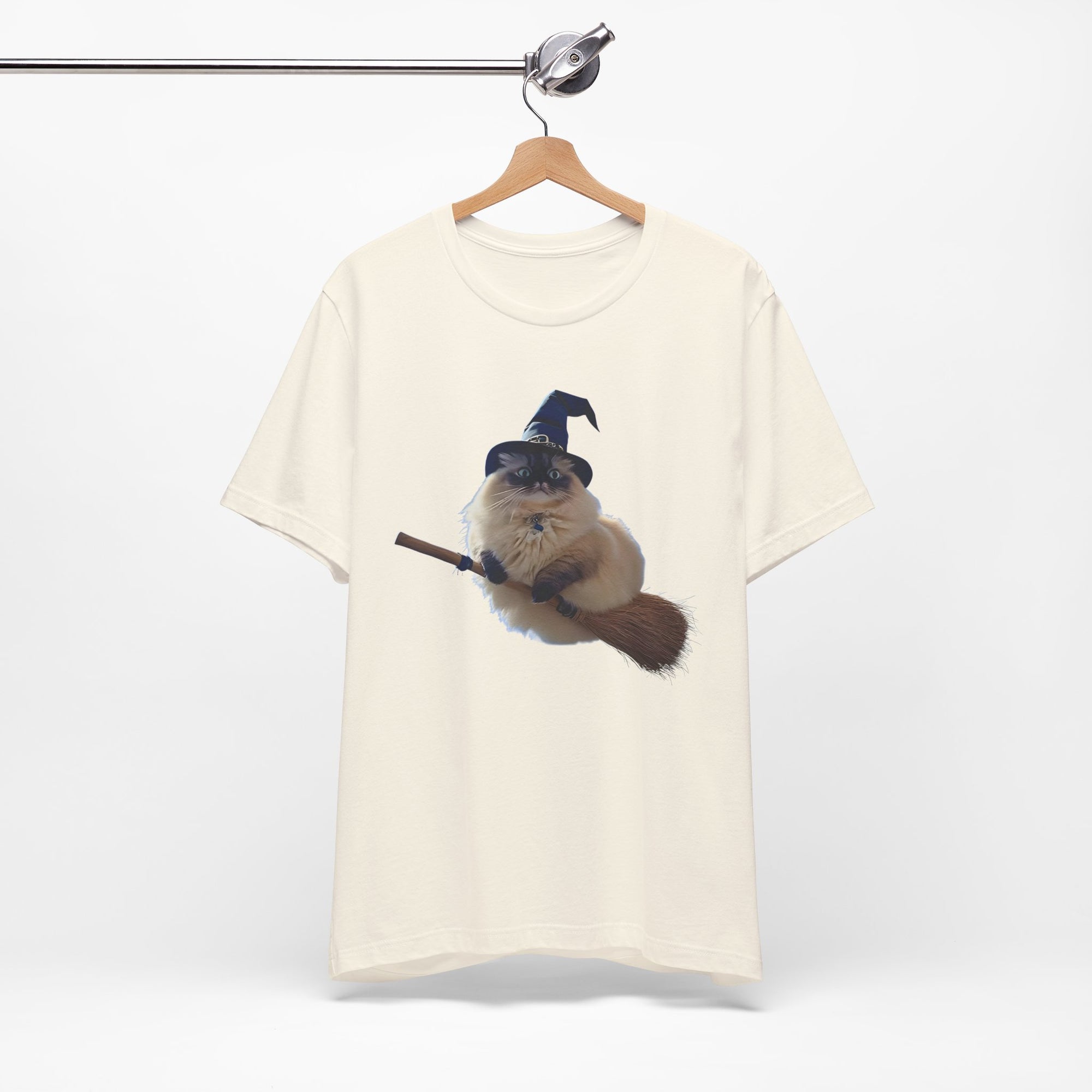 White t-shirt with a graphic of a cat wearing a witch hat and holding a broomstick, hanging on a wooden hanger against a white background.