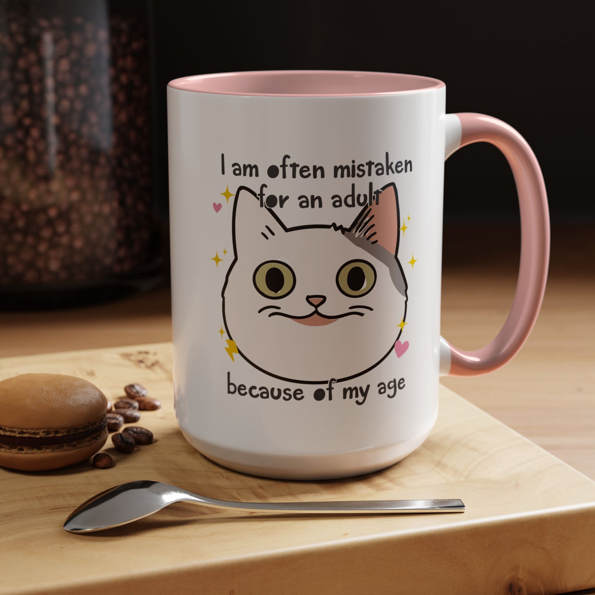 White ceramic mug with peach handle and interior featuring a cute wide-eyed cat face, hearts, sparkles, and the phrase “I am often mistaken for an adult because of my age.”

