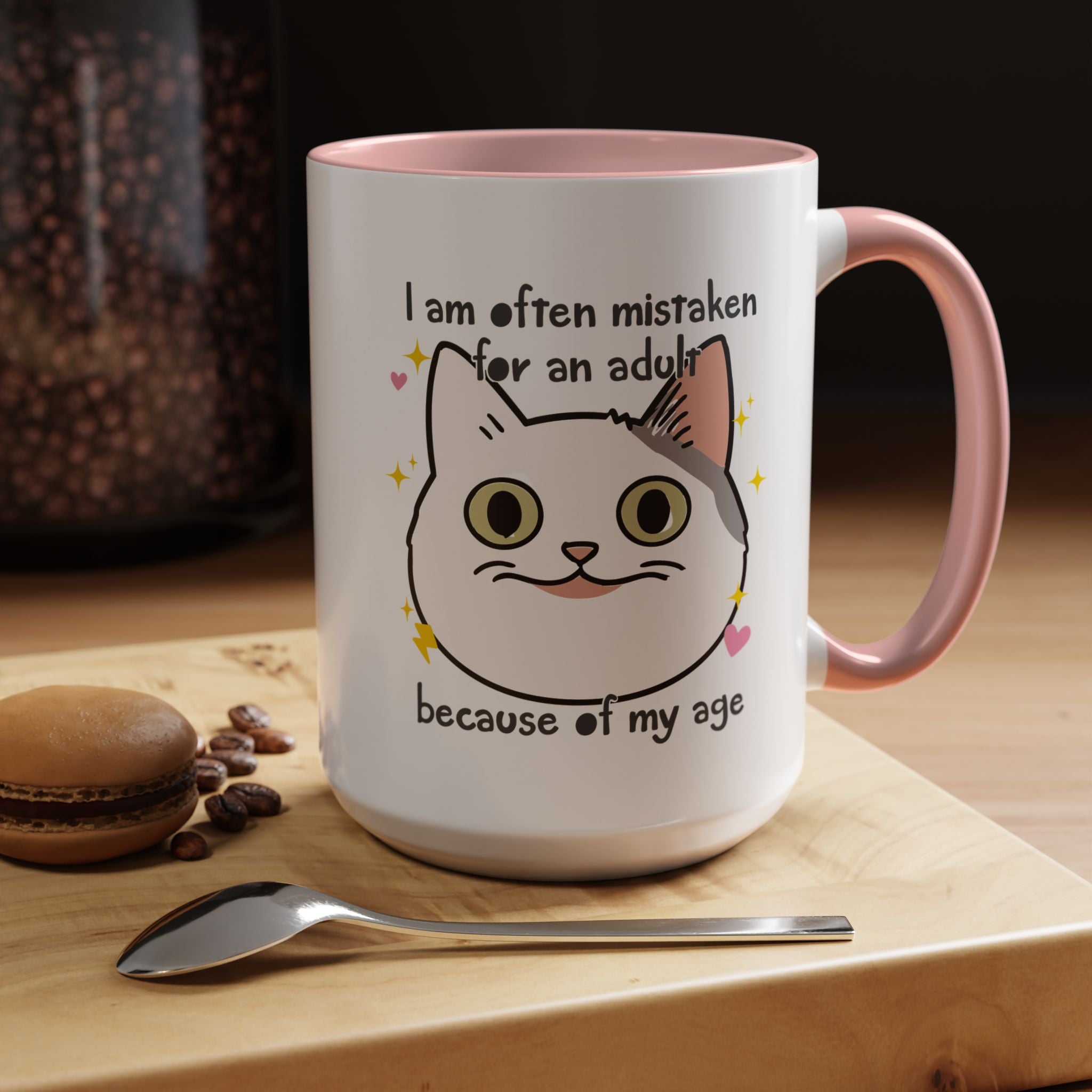 White ceramic mug with peach handle and interior featuring a cute wide-eyed cat face, hearts, sparkles, and the phrase “I am often mistaken for an adult because of my age.”
