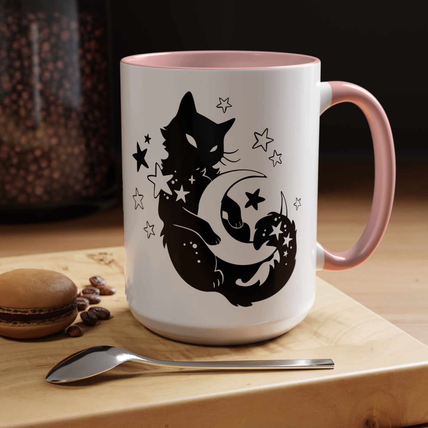 White mug with black cat and moon design on a wooden surface with coffee beans and a spoon.