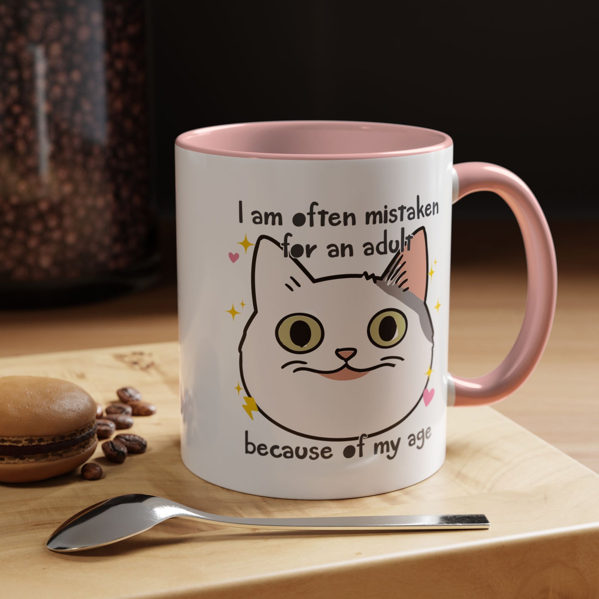 White ceramic mug with peach handle and interior featuring a cute wide-eyed cat face, hearts, sparkles, and the phrase “I am often mistaken for an adult because of my age.”
