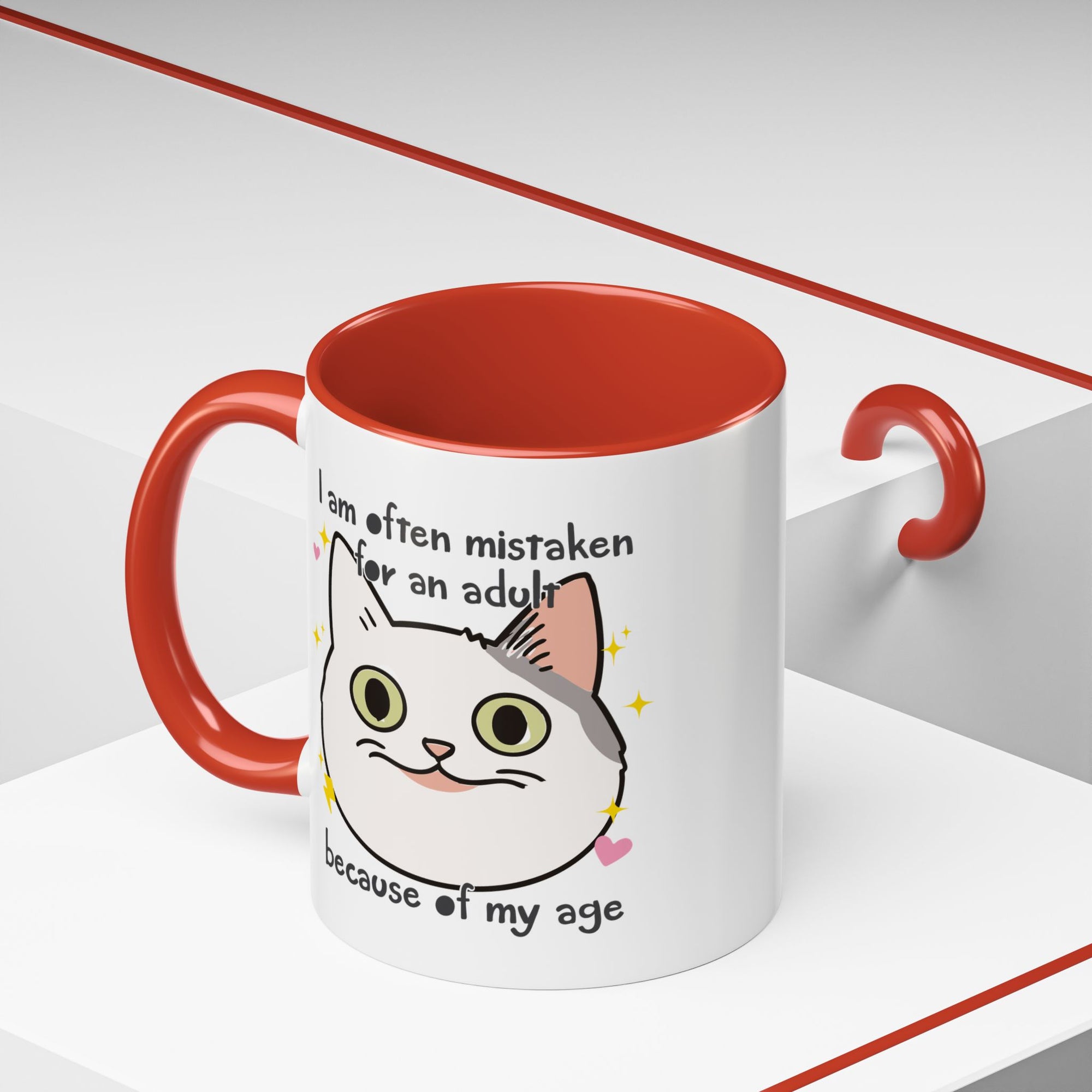 White ceramic mug with red handle and interior featuring a cute wide-eyed cat face, hearts, sparkles, and the phrase “I am often mistaken for an adult because of my age.”
