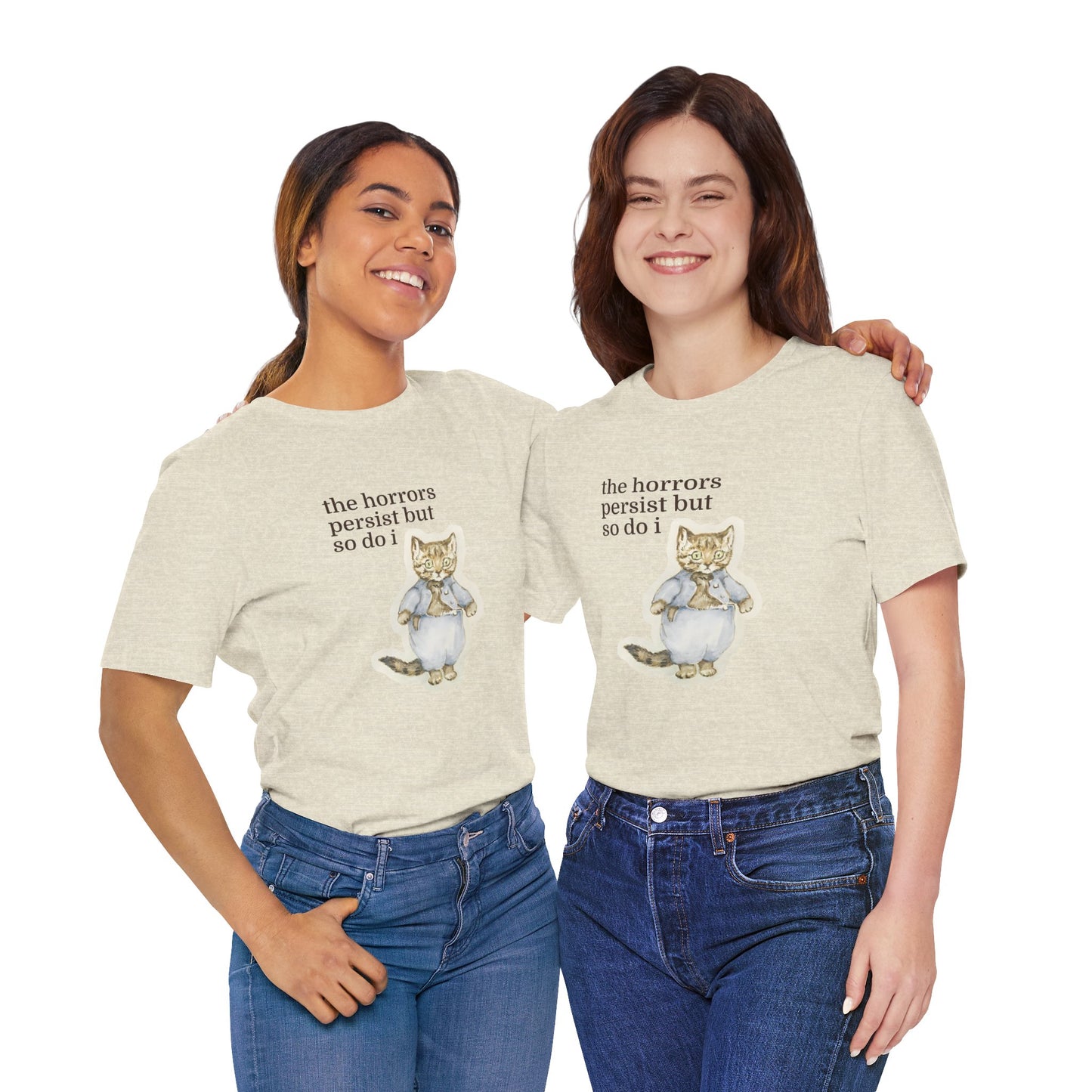 Two women wearing matching beige t-shirt featuring a vintage-style illustrated cat in blue clothing with the phrase “the horrors persist but so do i.”