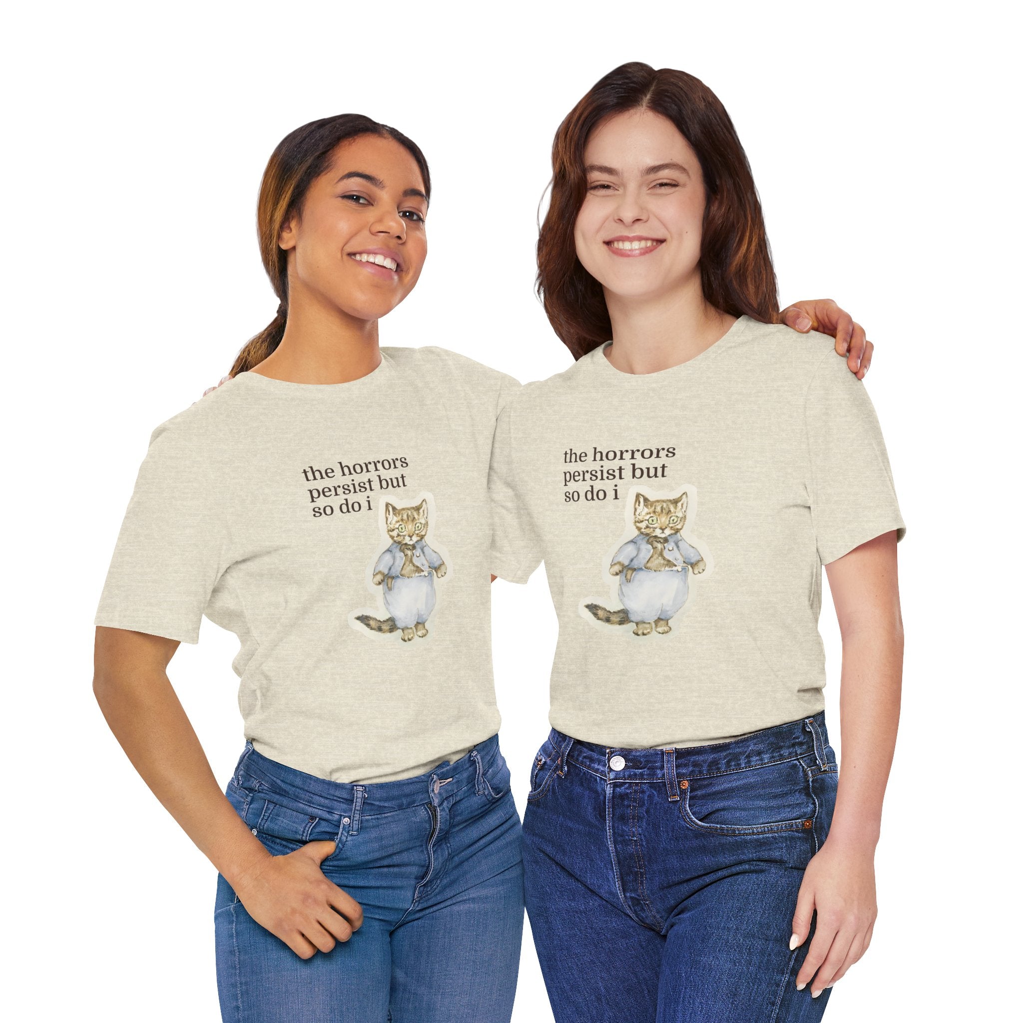 Two women wearing matching beige t-shirt featuring a vintage-style illustrated cat in blue clothing with the phrase “the horrors persist but so do i.”
