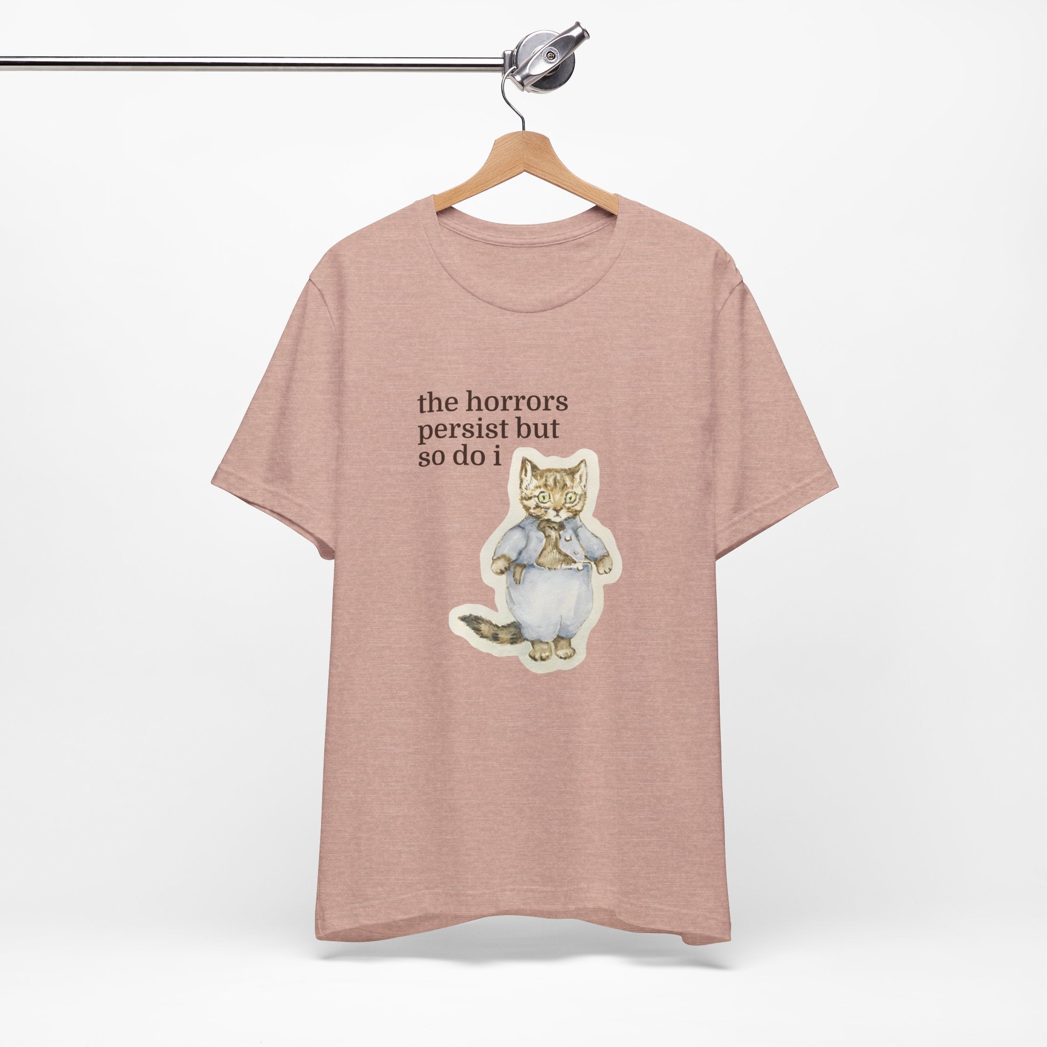 Peach t-shirt featuring a vintage-style illustrated cat in blue clothing with the phrase “the horrors persist but so do i.”