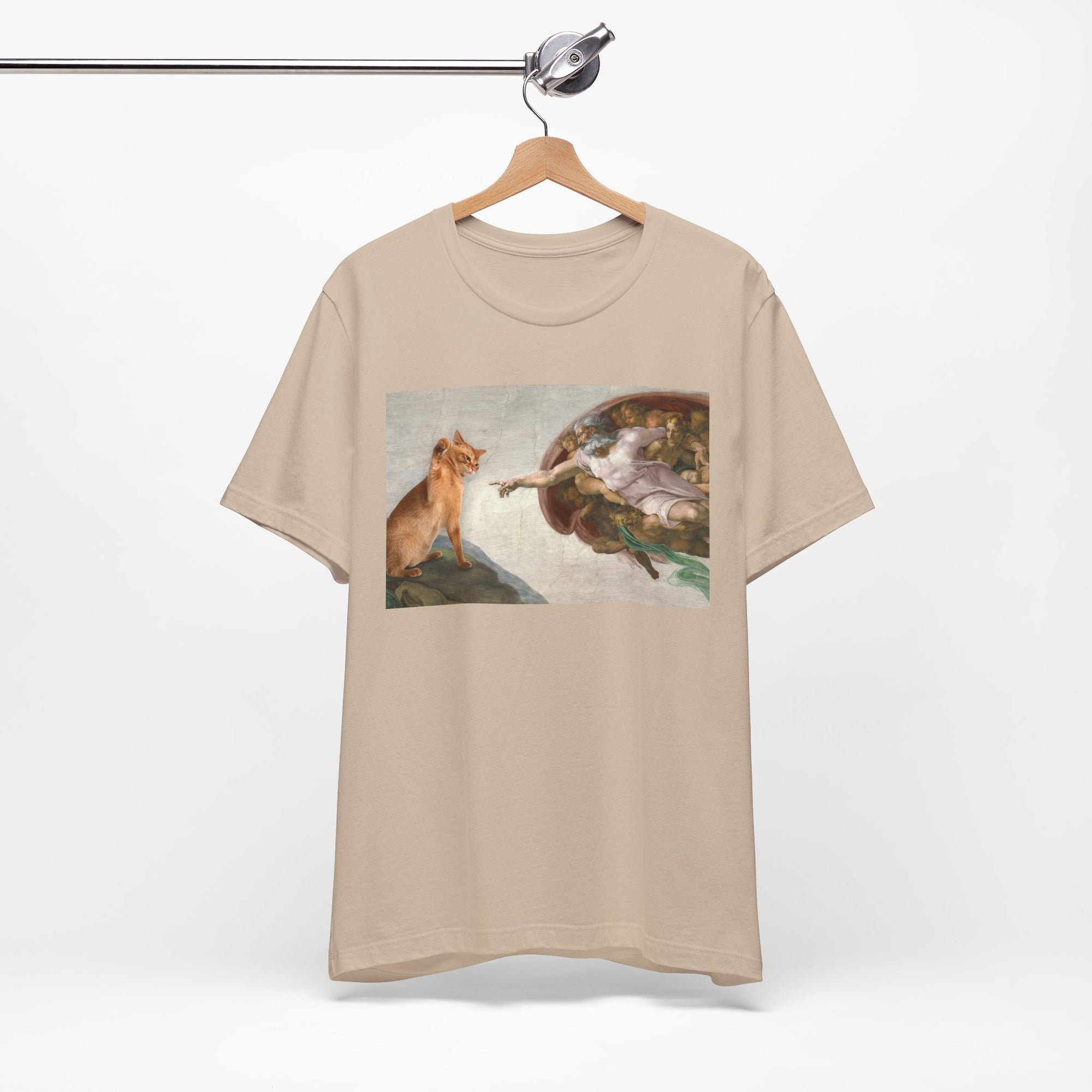 Beige t-shirt with a graphic design of a cat and god on a hanger against a white background