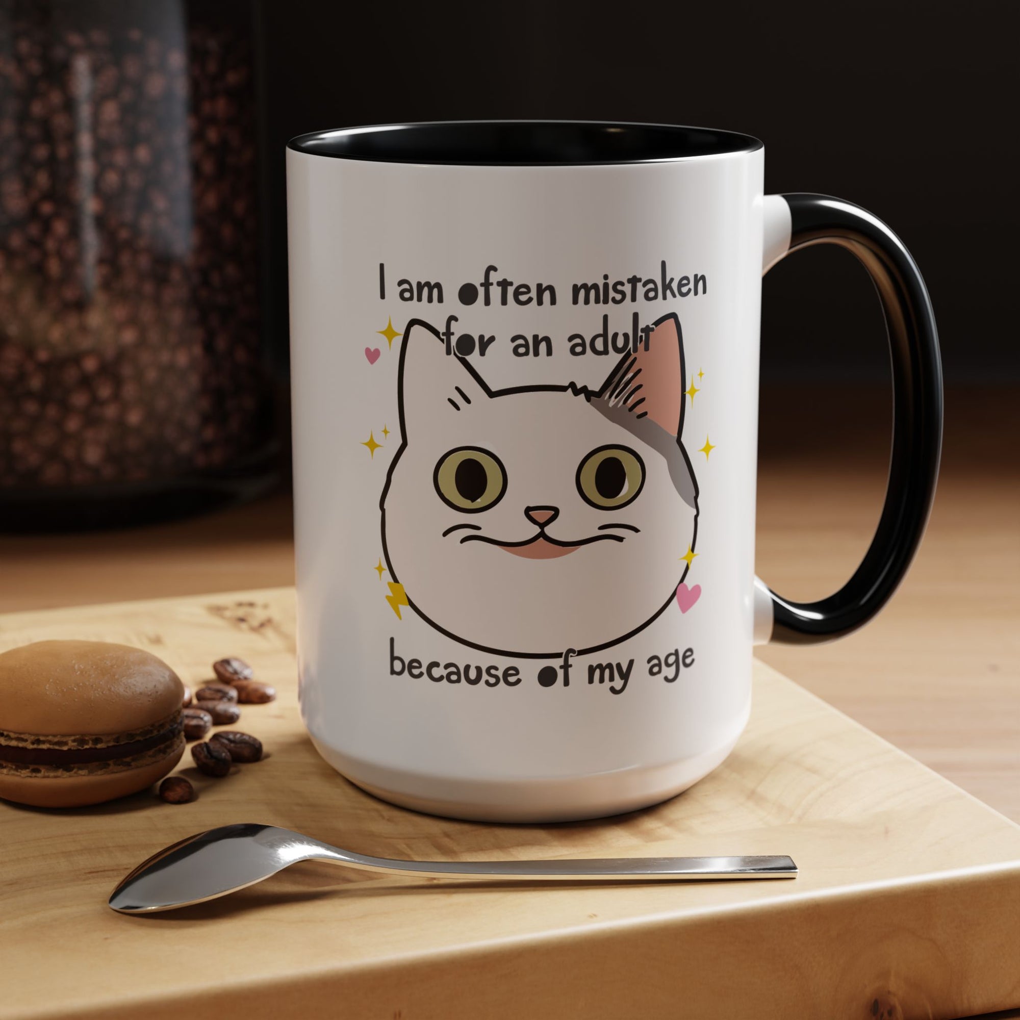 White ceramic mug with black handle and interior featuring a cute wide-eyed cat face, hearts, sparkles, and the phrase “I am often mistaken for an adult because of my age.”
