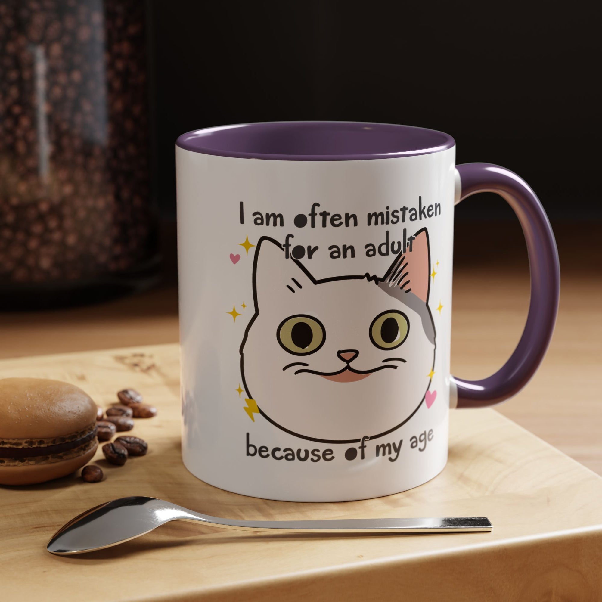 White ceramic mug with purple handle and interior featuring a cute wide-eyed cat face, hearts, sparkles, and the phrase “I am often mistaken for an adult because of my age.”
