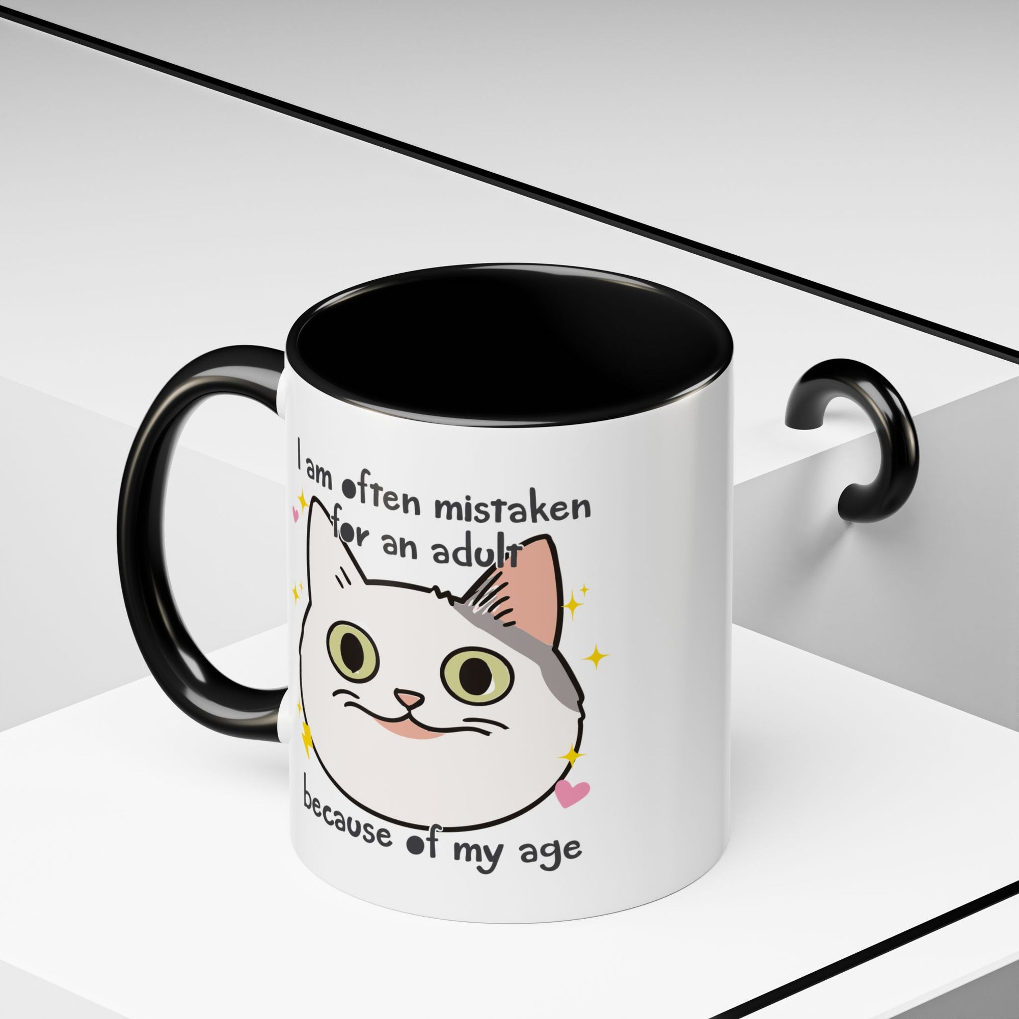 White ceramic mug with black handle and interior featuring a cute wide-eyed cat face, hearts, sparkles, and the phrase “I am often mistaken for an adult because of my age.”
