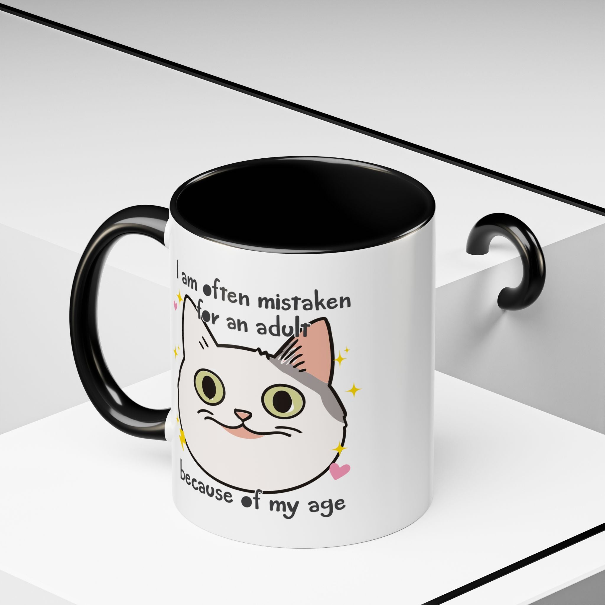 White ceramic mug with black handle and interior featuring a cute wide-eyed cat face, hearts, sparkles, and the phrase “I am often mistaken for an adult because of my age.”
