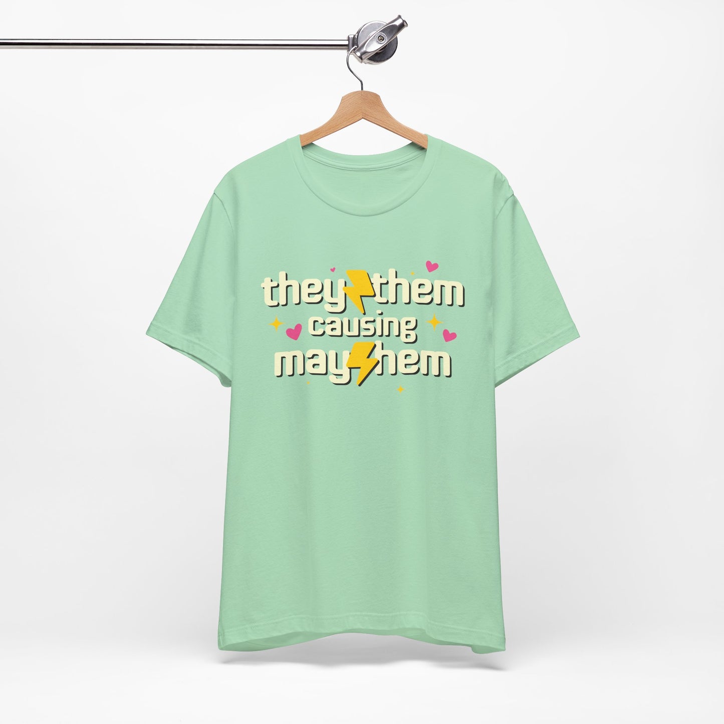 Mint green t-shirt with 'they/them causing mayhem' text on a hanger against a white background