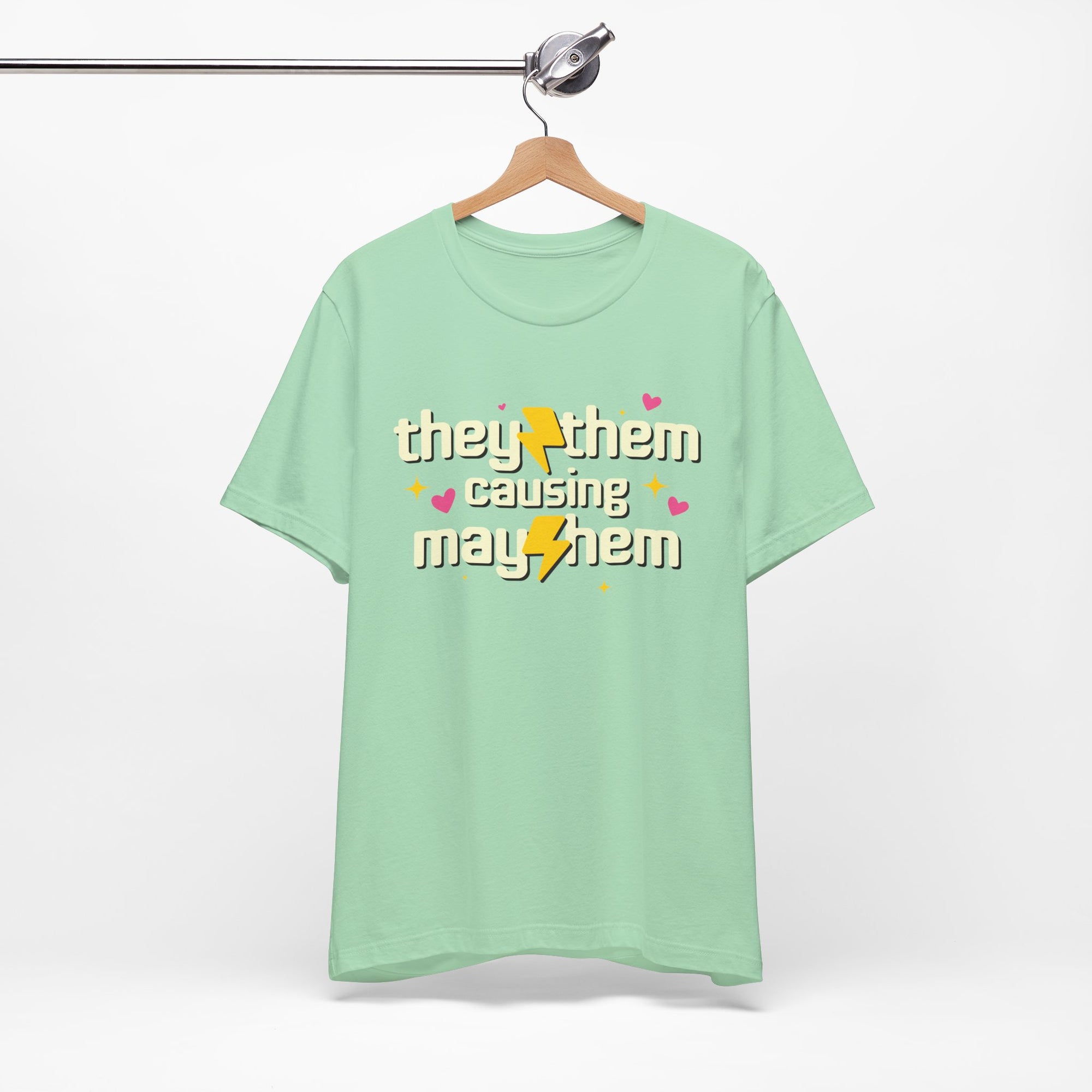 Mint green t-shirt with 'they/them causing mayhem' text on a hanger against a white background