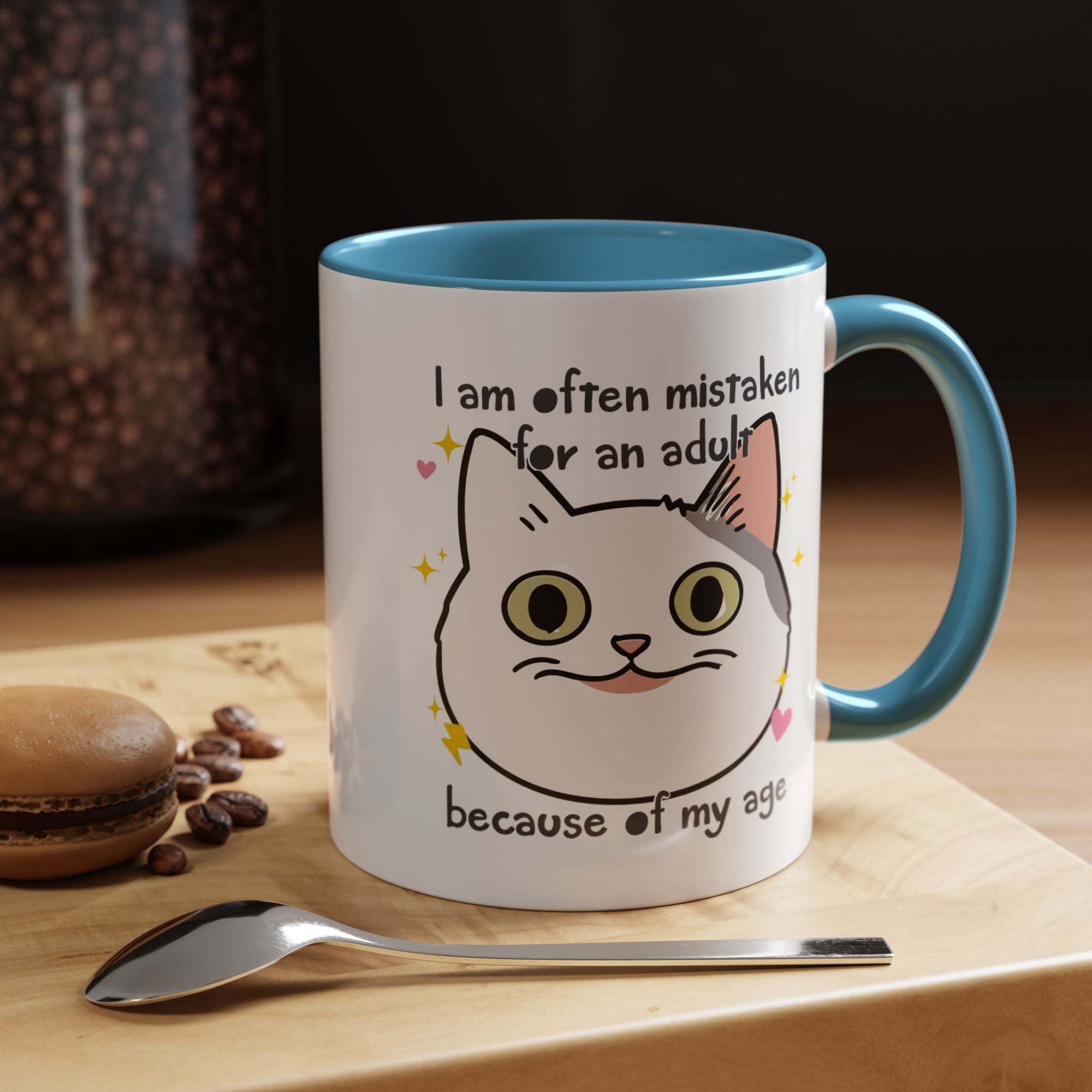 White ceramic mug with blue handle and interior featuring a cute wide-eyed cat face, hearts, sparkles, and the phrase “I am often mistaken for an adult because of my age.”

