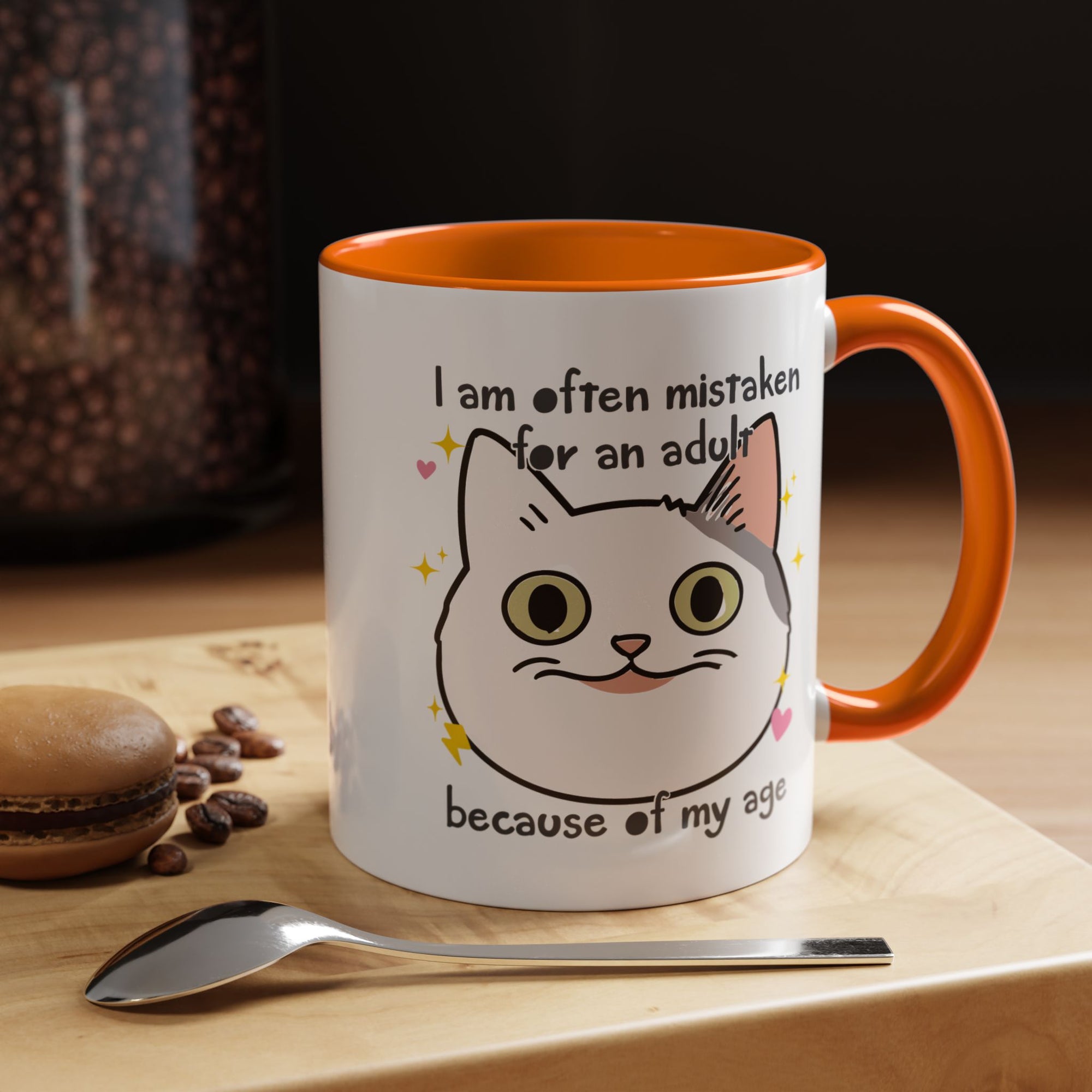 White ceramic mug with orange handle and interior featuring a cute wide-eyed cat face, hearts, sparkles, and the phrase “I am often mistaken for an adult because of my age.”
