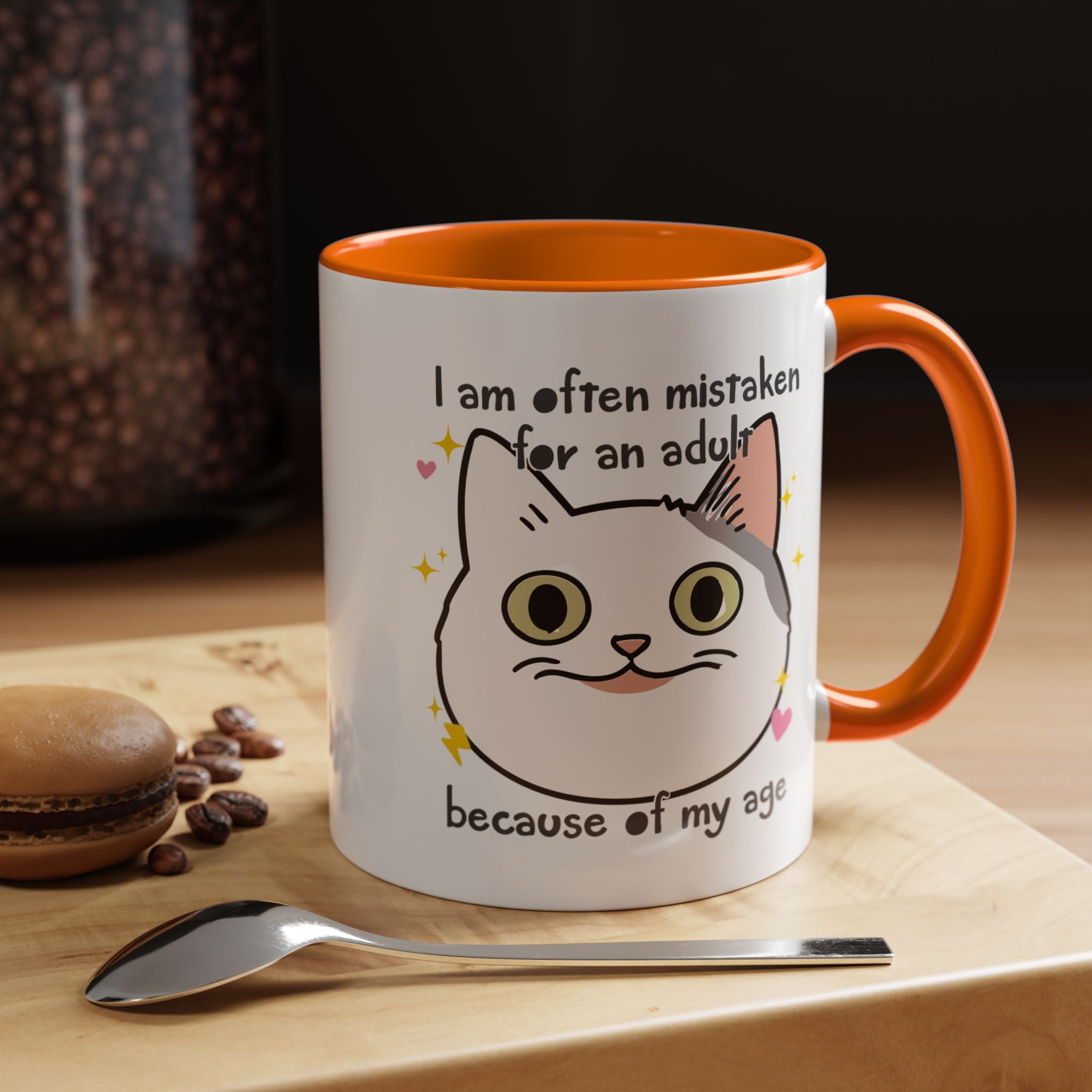 White ceramic mug with orange handle and interior featuring a cute wide-eyed cat face, hearts, sparkles, and the phrase “I am often mistaken for an adult because of my age.”
