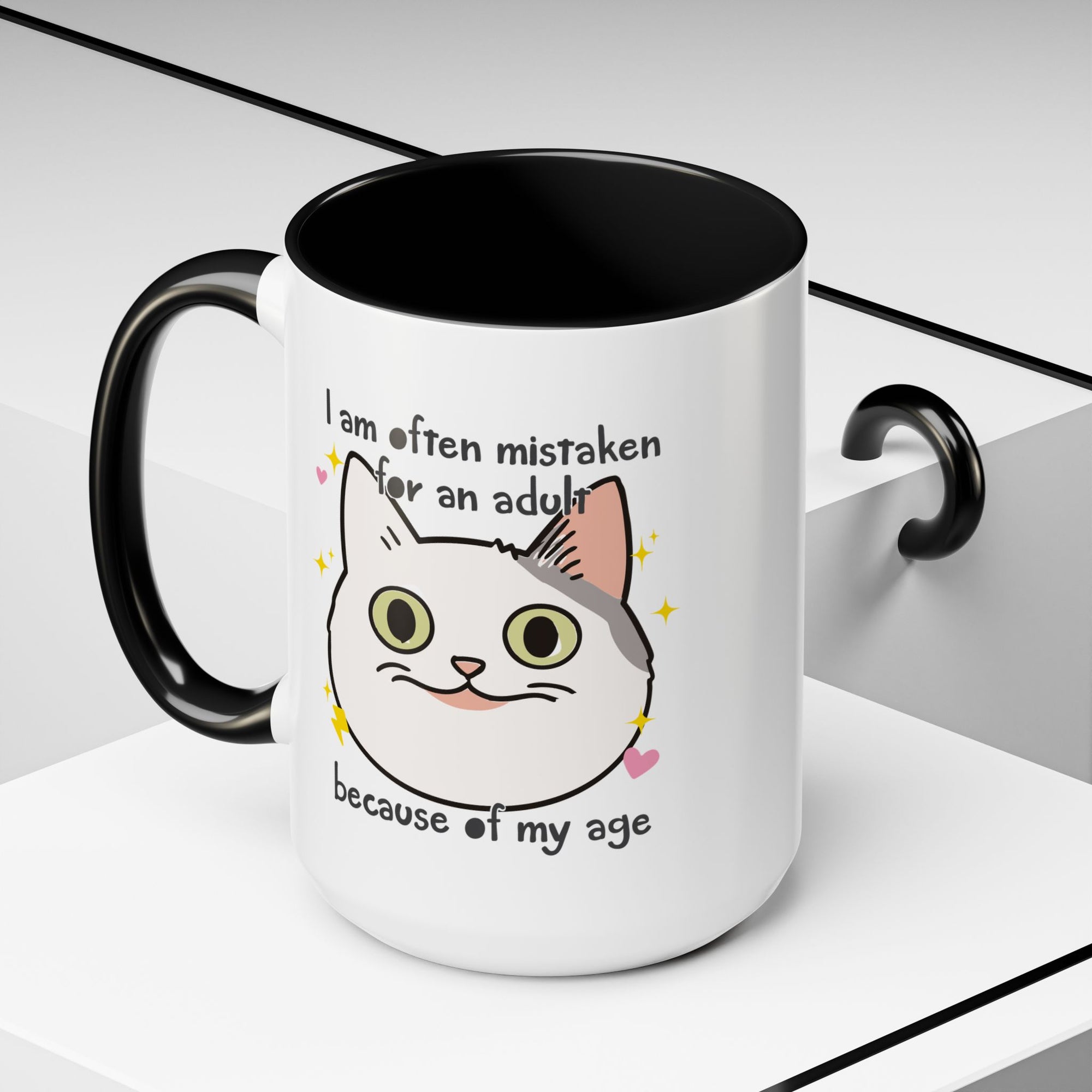 White ceramic mug with black handle and interior featuring a cute wide-eyed cat face, hearts, sparkles, and the phrase “I am often mistaken for an adult because of my age.”
