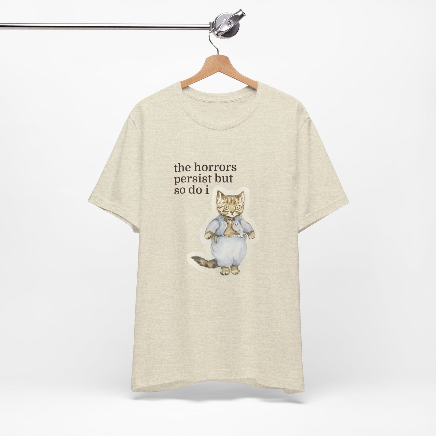 Beige t-shirt featuring a vintage-style illustrated cat in blue clothing with the phrase “the horrors persist but so do i.”