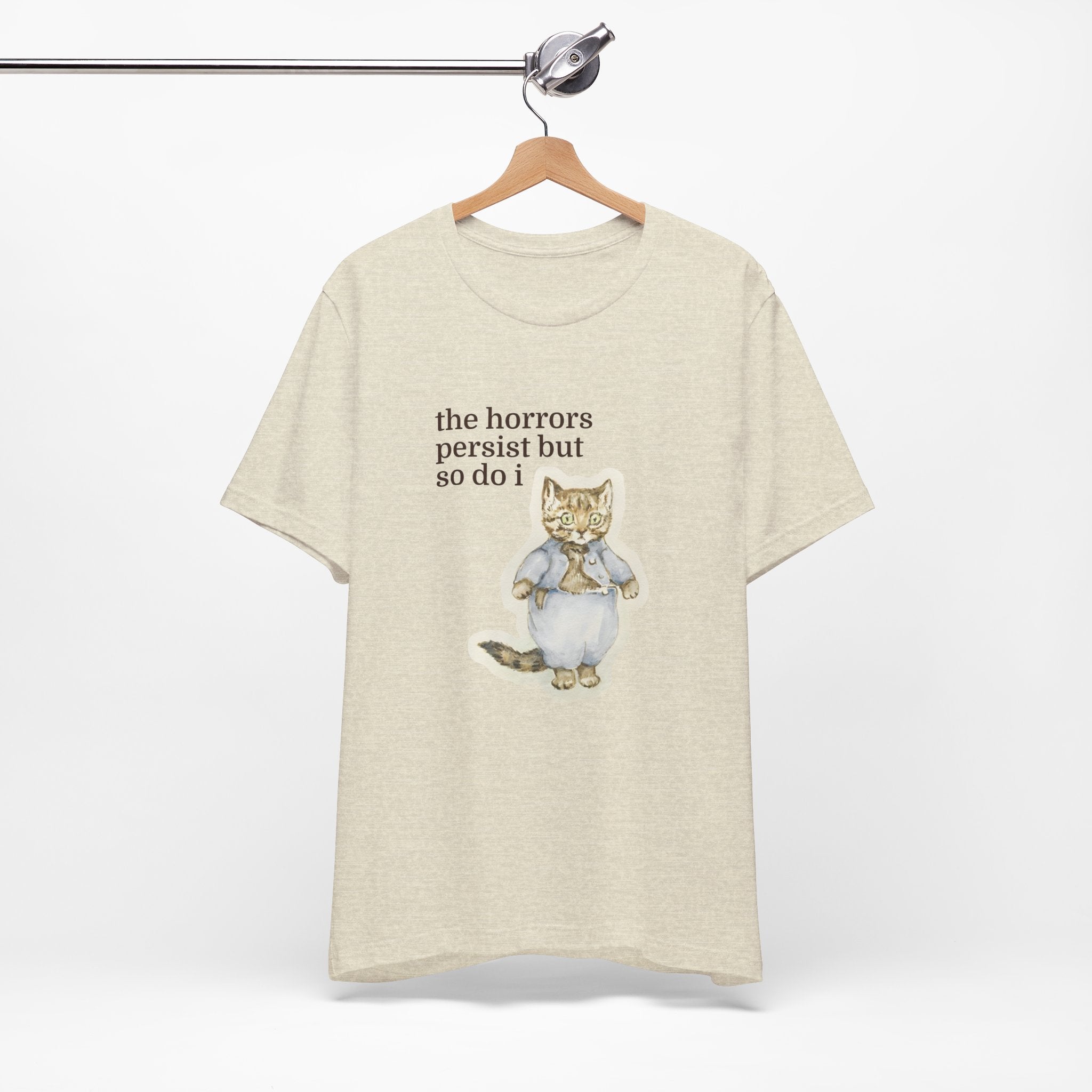 Beige t-shirt featuring a vintage-style illustrated cat in blue clothing with the phrase “the horrors persist but so do i.”