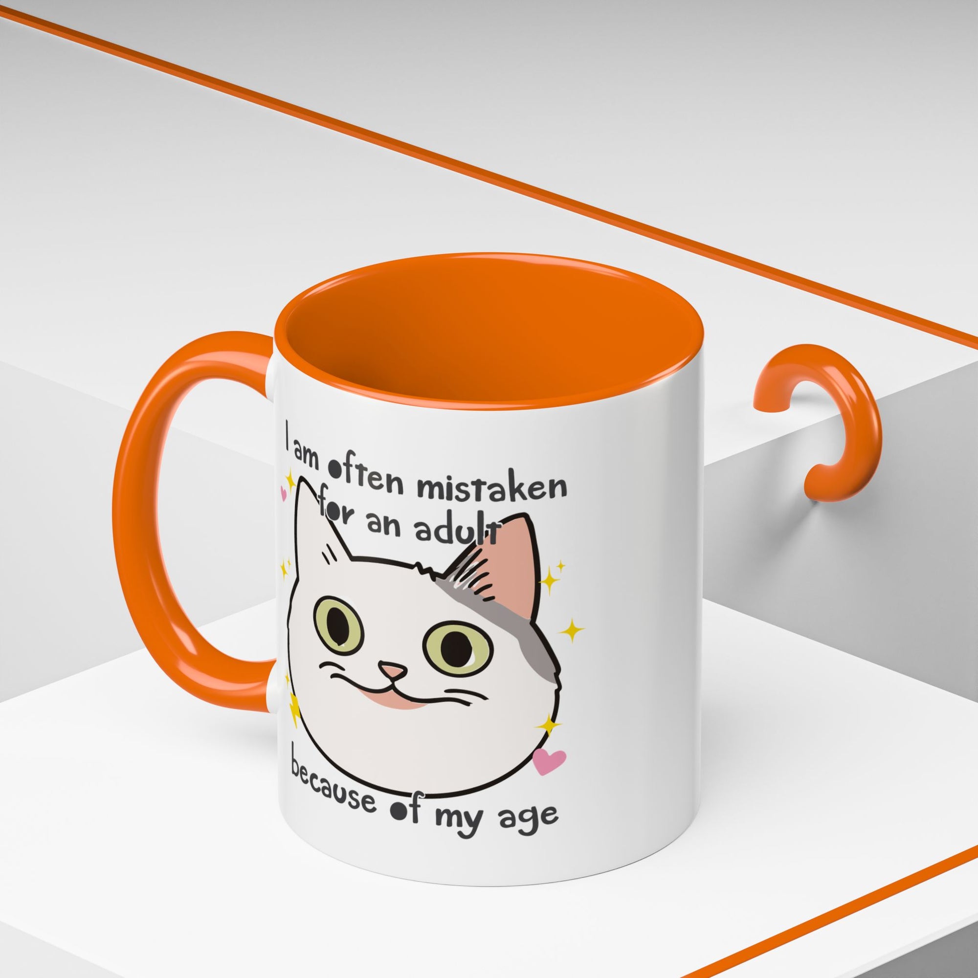 White ceramic mug with orange handle and interior featuring a cute wide-eyed cat face, hearts, sparkles, and the phrase “I am often mistaken for an adult because of my age.”

