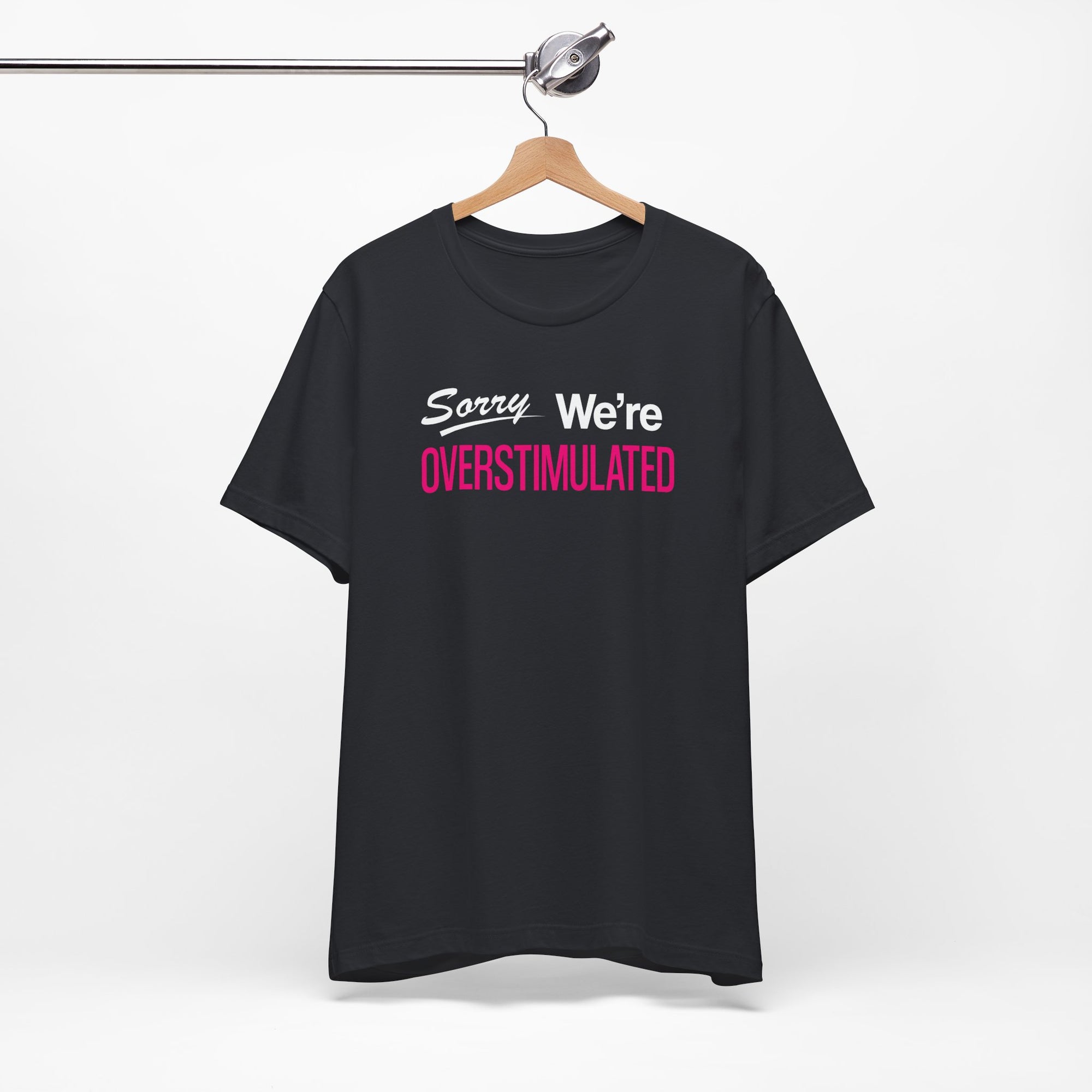 Black t-shirt featuring the phrase “Sorry We’re Overstimulated” in white and hot pink text.
