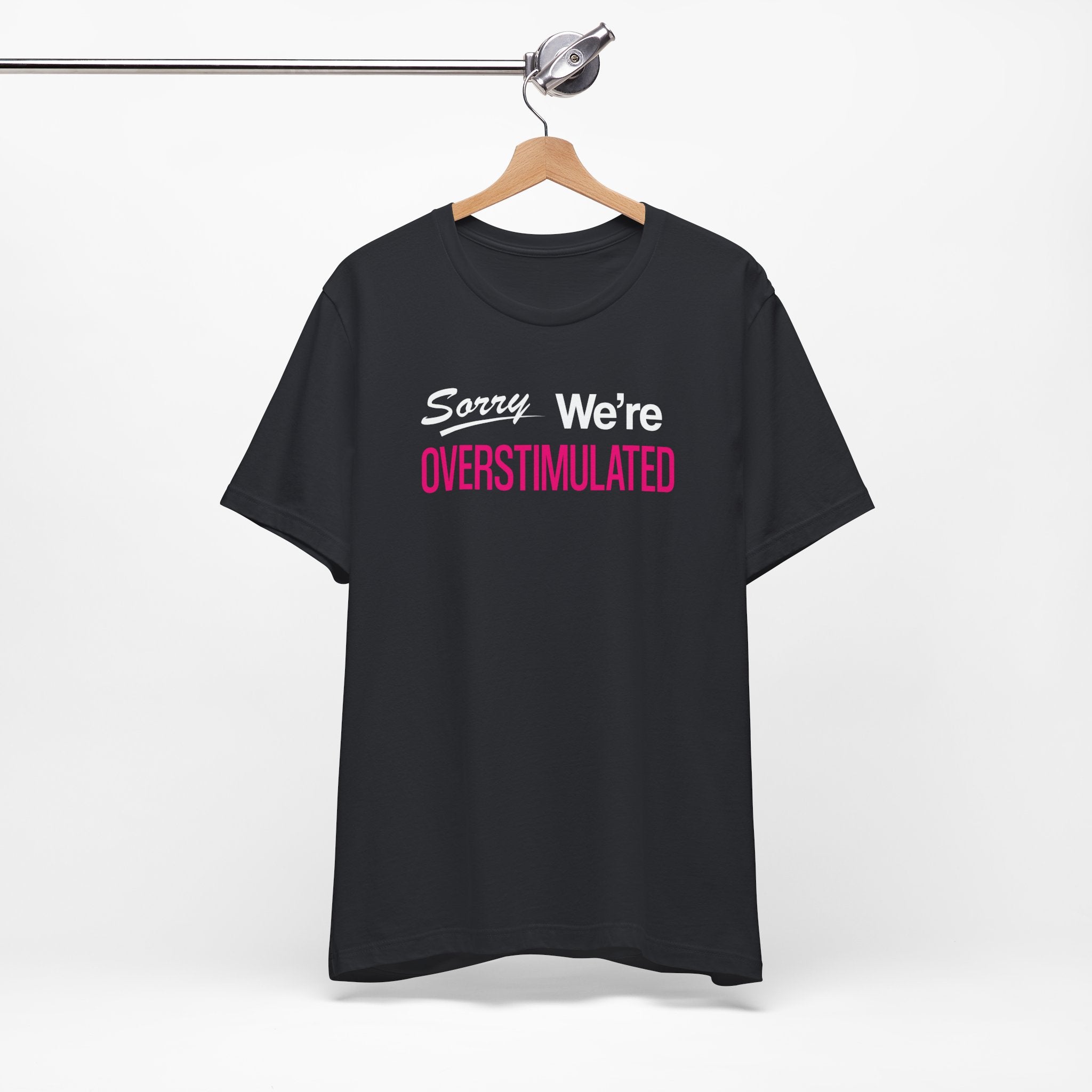 Black t-shirt featuring the phrase “Sorry We’re Overstimulated” in white and hot pink text.
