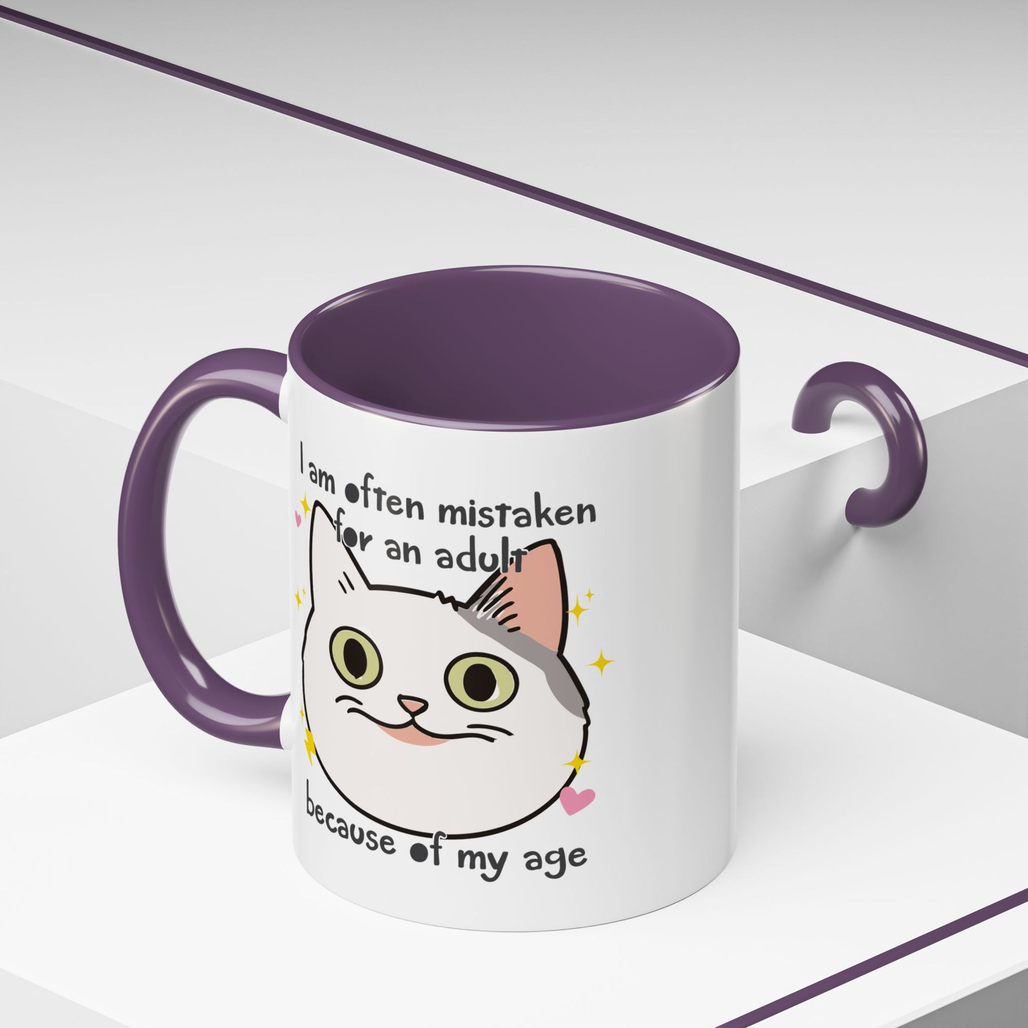 White ceramic mug with purple handle and interior featuring a cute wide-eyed cat face, hearts, sparkles, and the phrase “I am often mistaken for an adult because of my age.”
