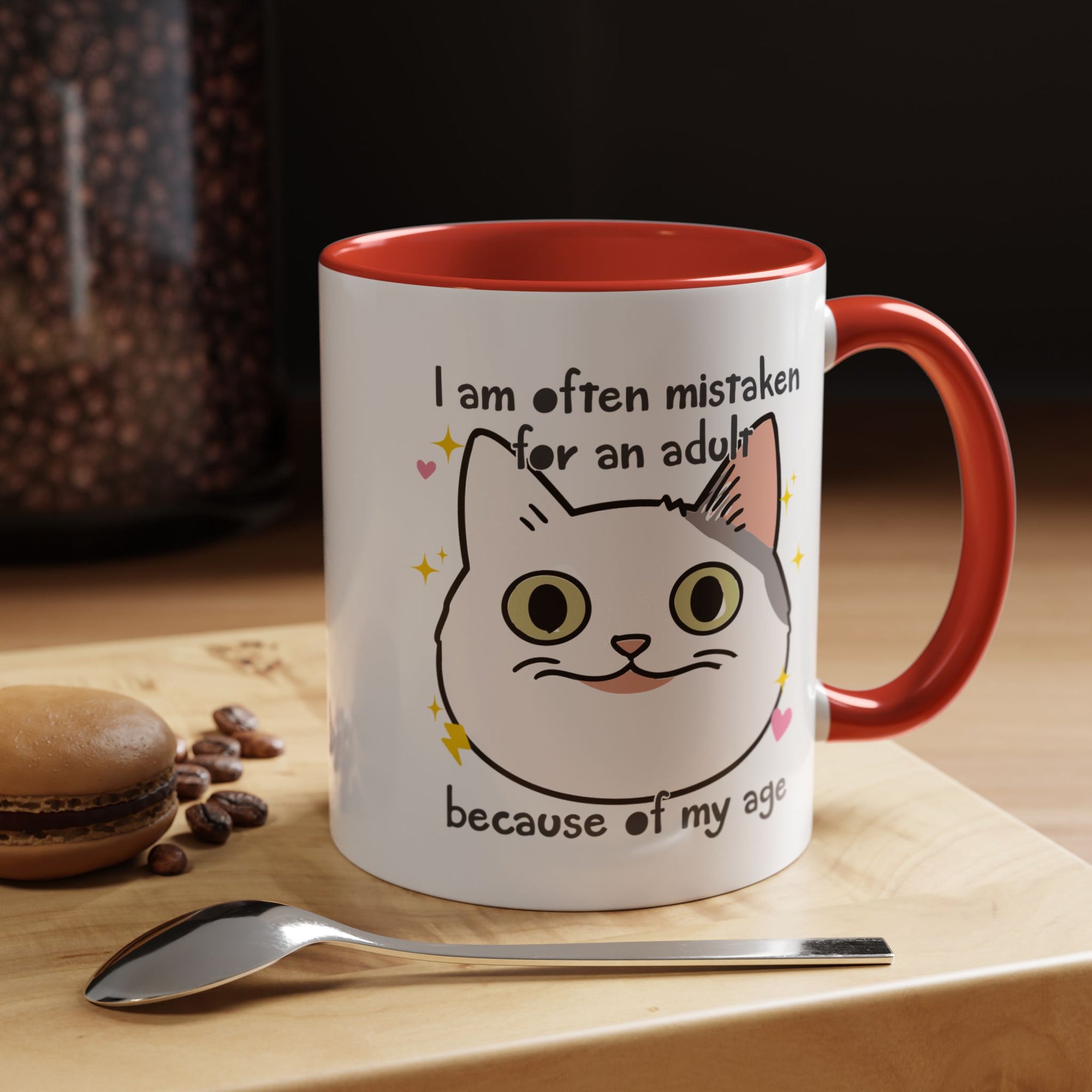 White ceramic mug with red handle and interior featuring a cute wide-eyed cat face, hearts, sparkles, and the phrase “I am often mistaken for an adult because of my age.”

