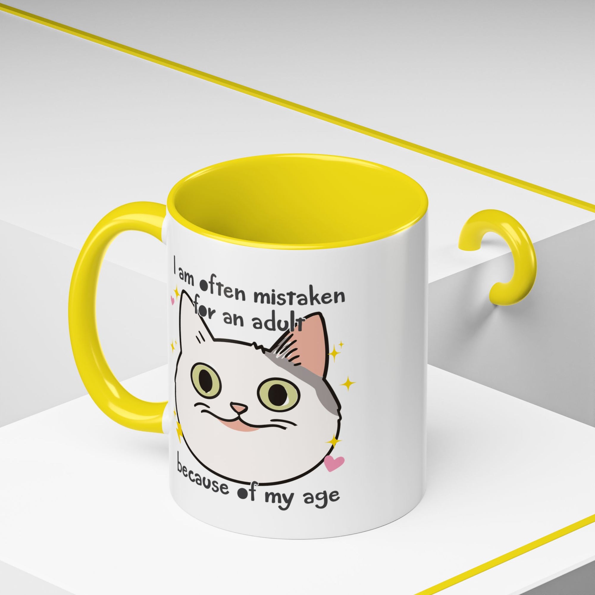 White ceramic mug with yellow handle and interior featuring a cute wide-eyed cat face, hearts, sparkles, and the phrase “I am often mistaken for an adult because of my age.”
