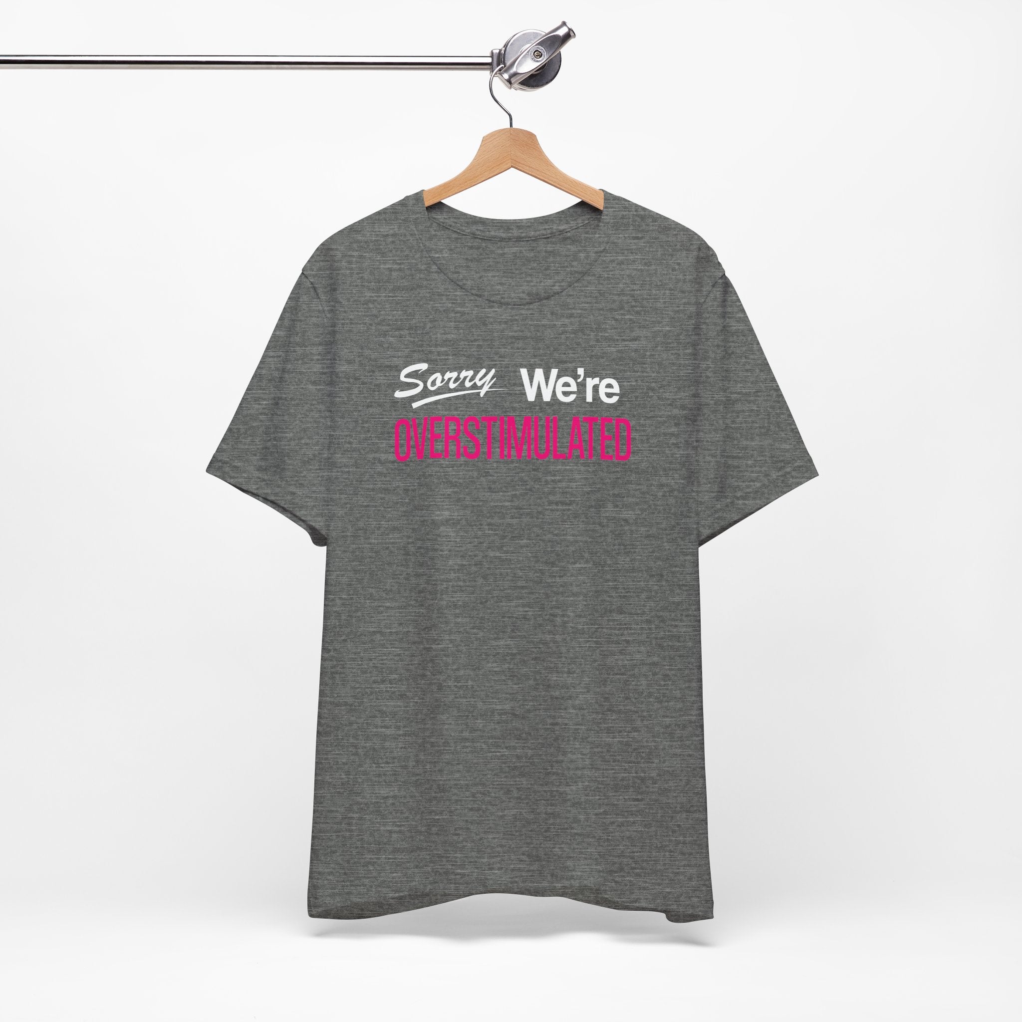 Grey t-shirt featuring the phrase “Sorry We’re Overstimulated” in white and hot pink text.
