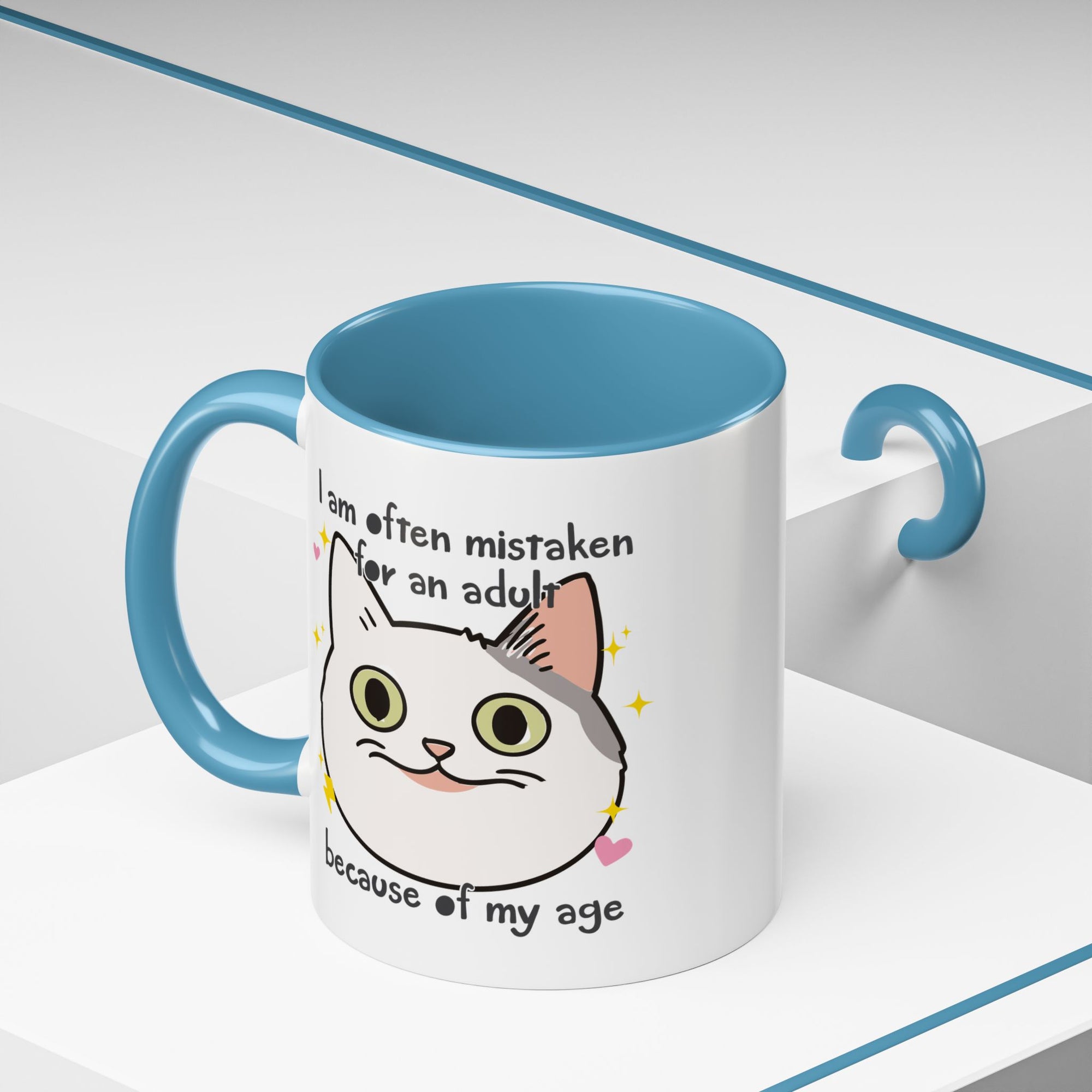 White ceramic mug with blue handle and interior featuring a cute wide-eyed cat face, hearts, sparkles, and the phrase “I am often mistaken for an adult because of my age.”

