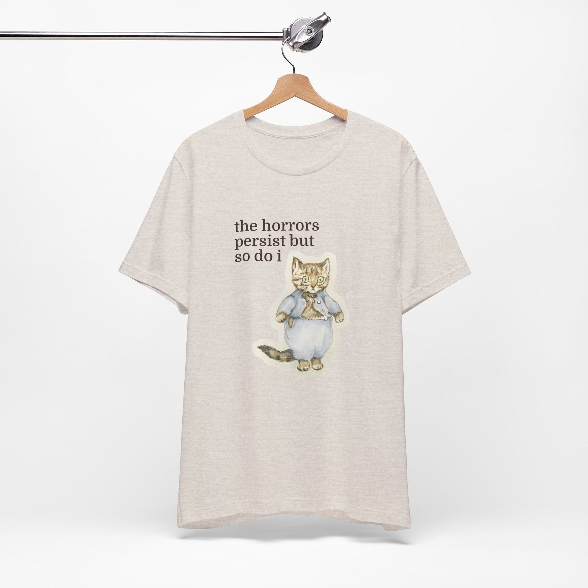 Beige t-shirt featuring a vintage-style illustrated cat in blue clothing with the phrase “the horrors persist but so do i.”