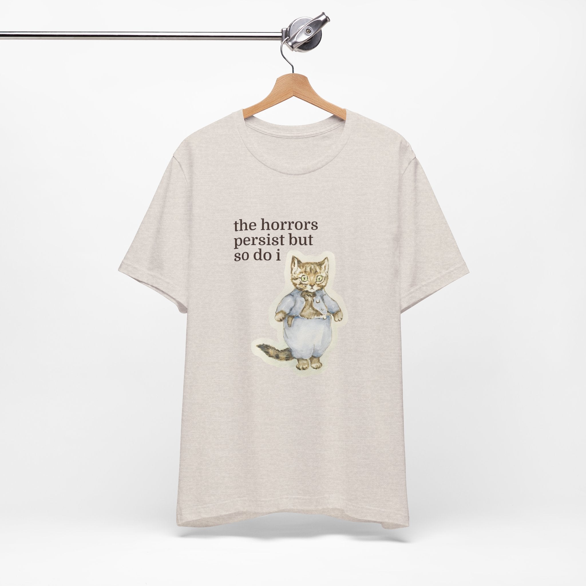 Beige t-shirt featuring a vintage-style illustrated cat in blue clothing with the phrase “the horrors persist but so do i.”