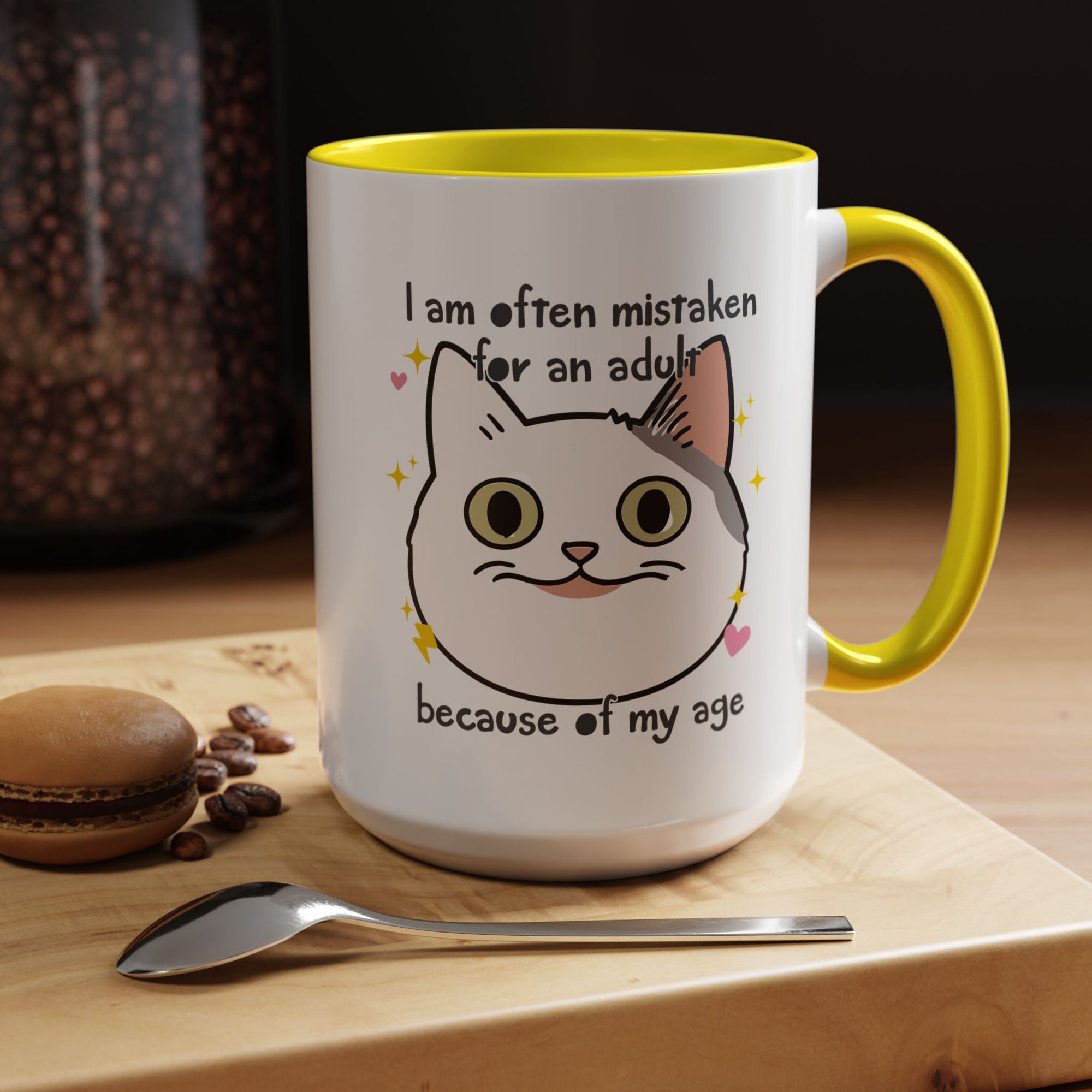 White ceramic mug with yellow handle and interior featuring a cute wide-eyed cat face, hearts, sparkles, and the phrase “I am often mistaken for an adult because of my age.”
