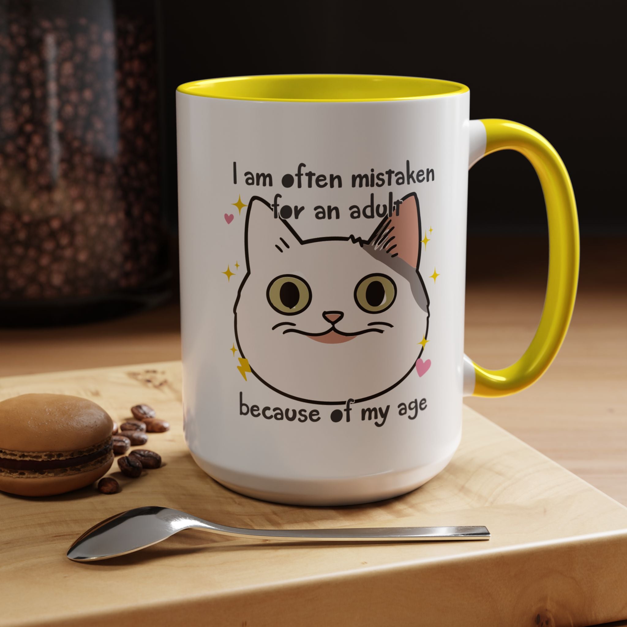 White ceramic mug with yellow handle and interior featuring a cute wide-eyed cat face, hearts, sparkles, and the phrase “I am often mistaken for an adult because of my age.”
