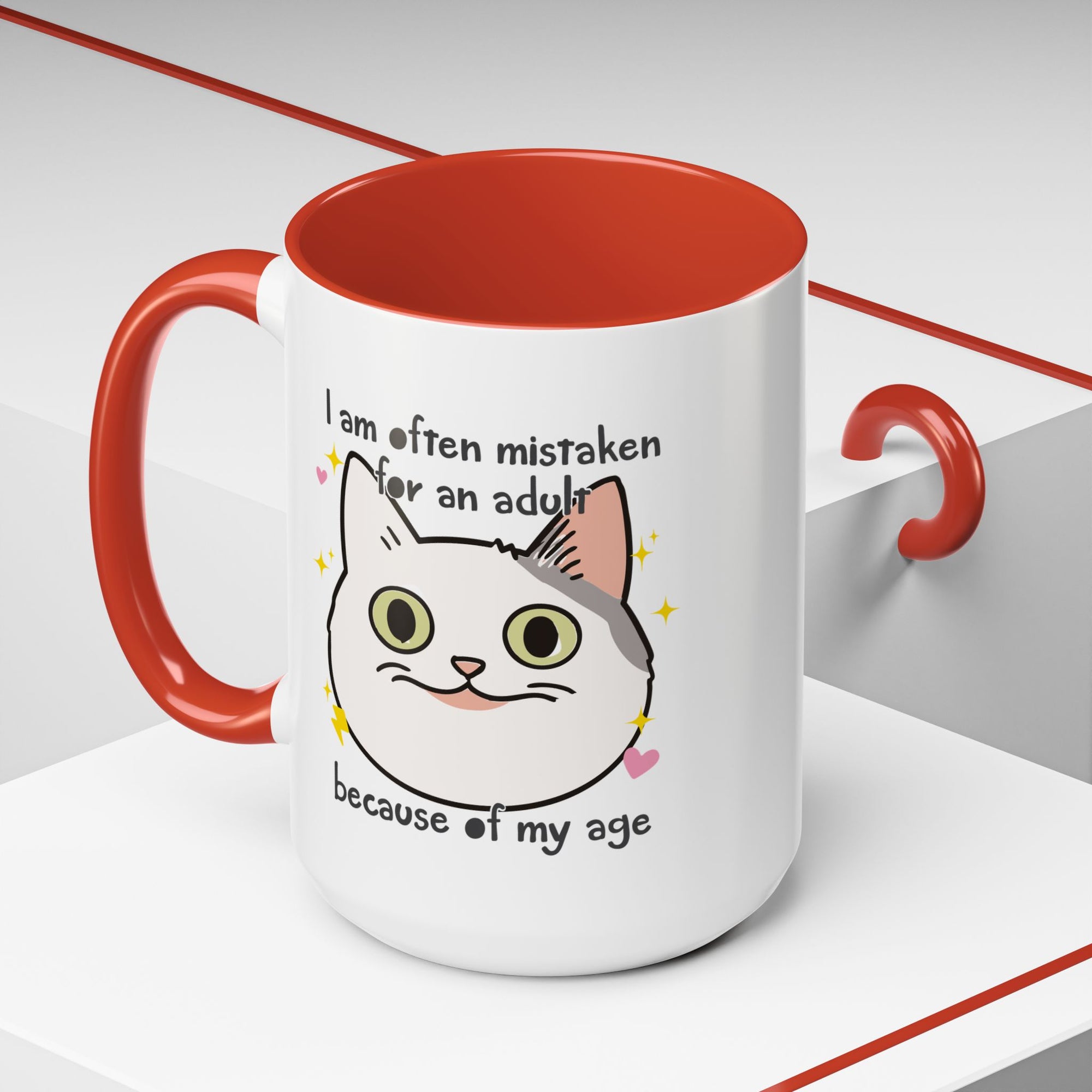 White ceramic mug with red handle and interior featuring a cute wide-eyed cat face, hearts, sparkles, and the phrase “I am often mistaken for an adult because of my age.”
