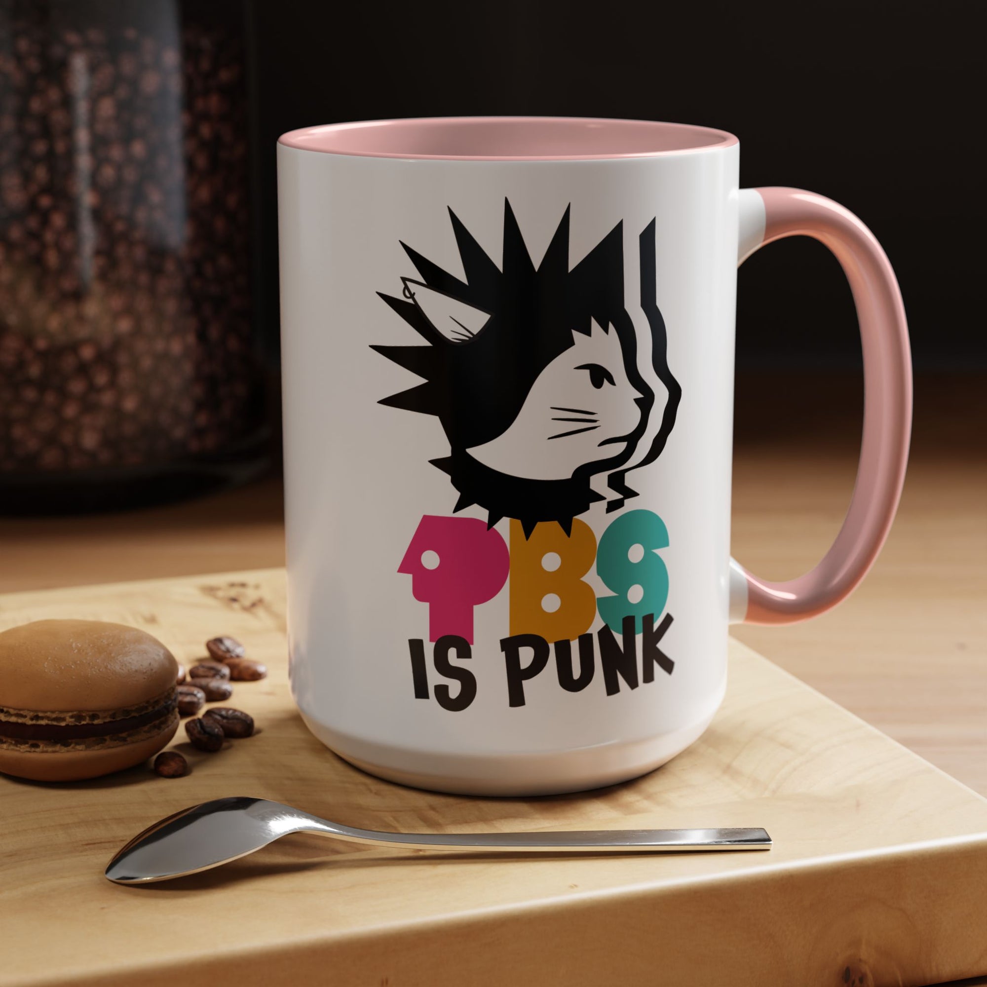 White mug with pink handle featuring a cat graphic and 'PBS is Punk' text on a wooden surface with coffee beans and a macaron.