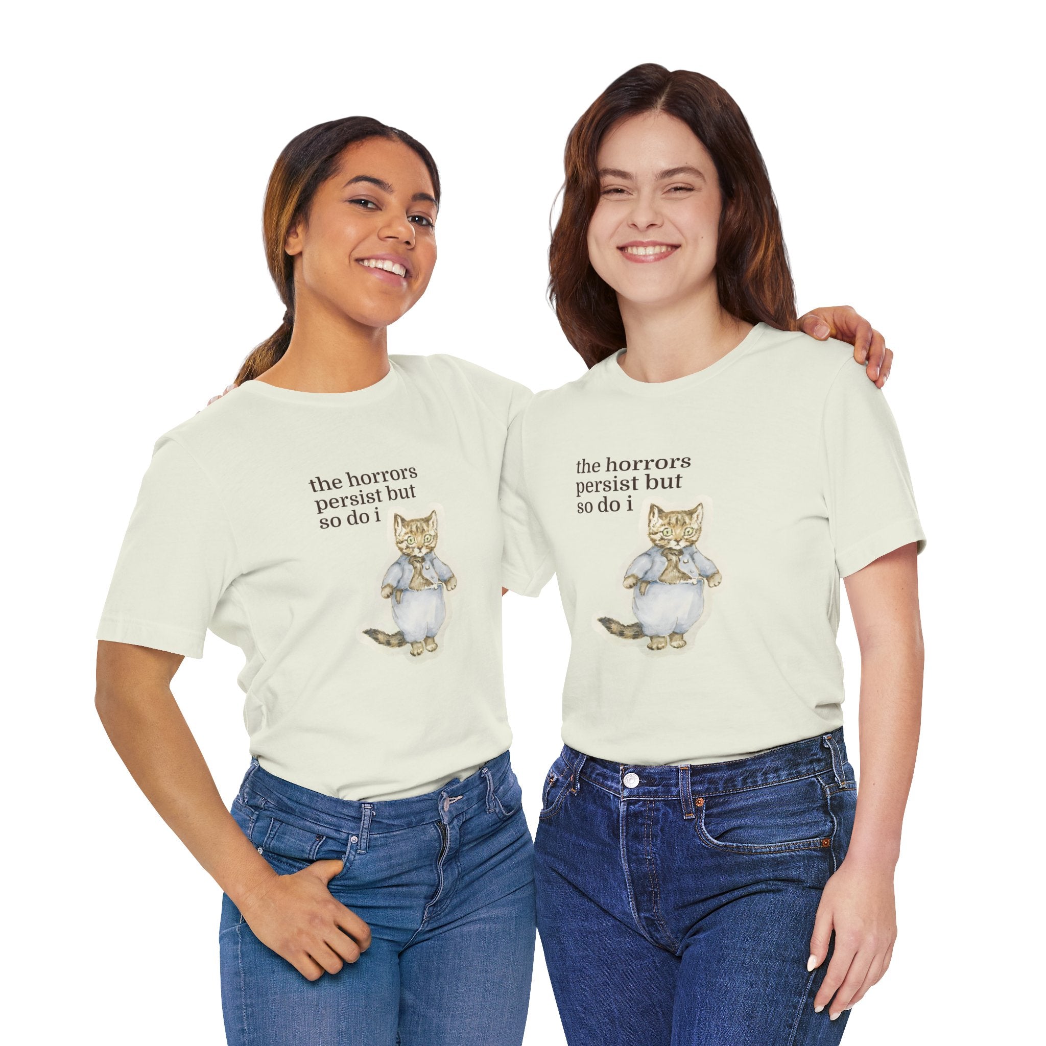 Two women wearing matching beige t-shirt featuring a vintage-style illustrated cat in blue clothing with the phrase “the horrors persist but so do i.”