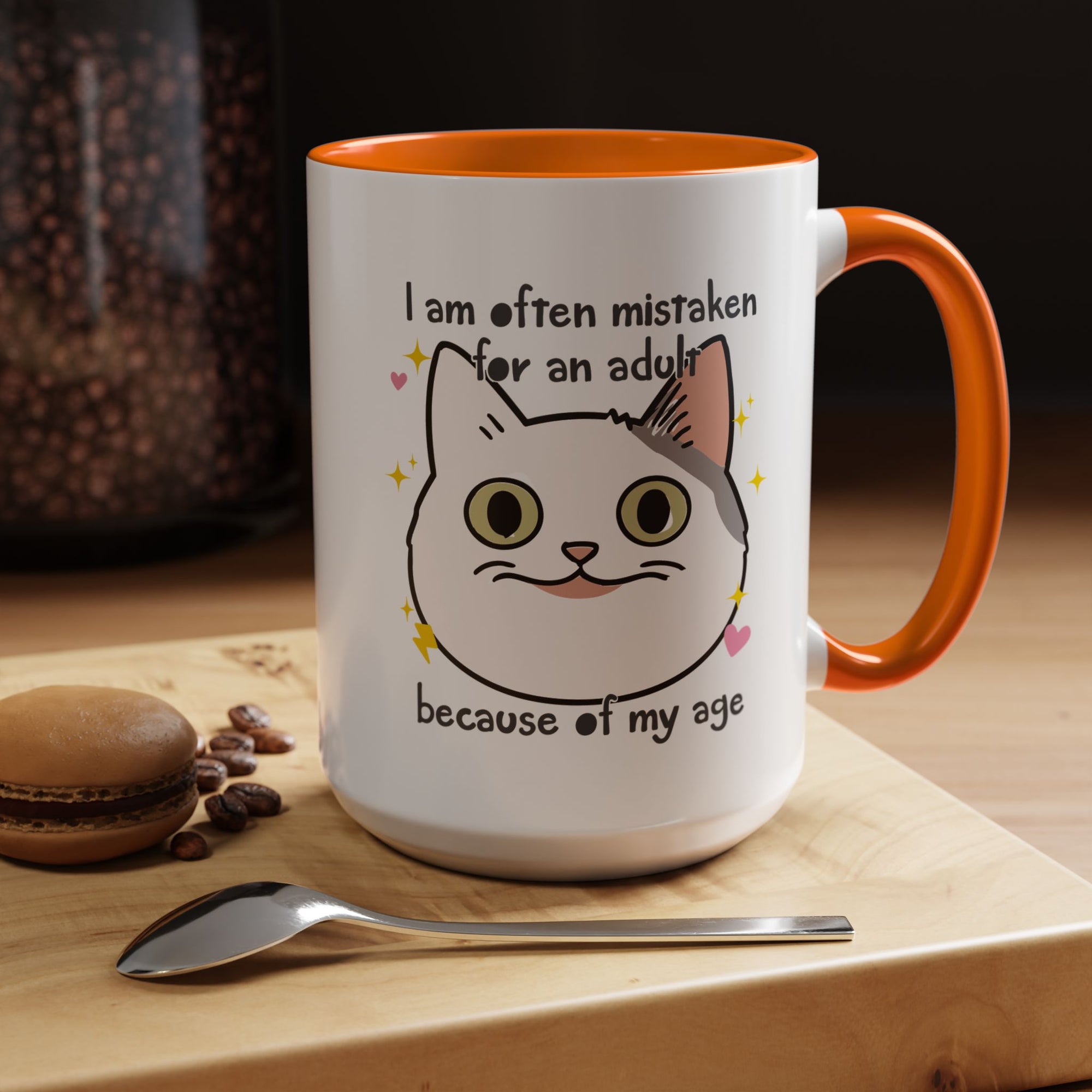 White ceramic mug with orange handle and interior featuring a cute wide-eyed cat face, hearts, sparkles, and the phrase “I am often mistaken for an adult because of my age.”
