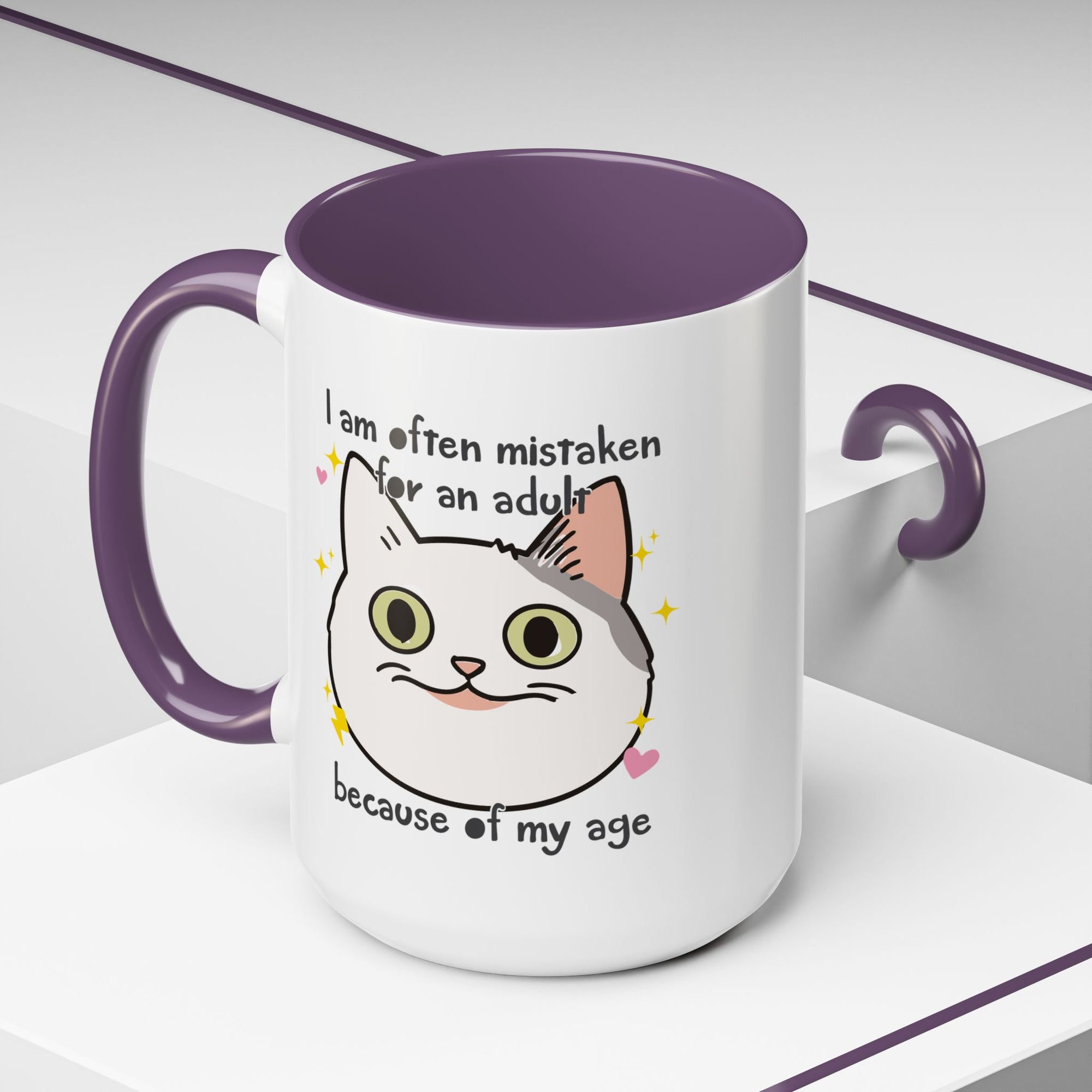 White ceramic mug with purple handle and interior featuring a cute wide-eyed cat face, hearts, sparkles, and the phrase “I am often mistaken for an adult because of my age.”
