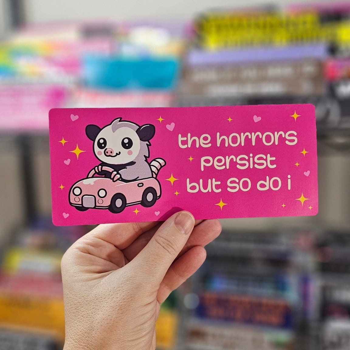 rectangular bumper stickers that say “the horrors persist but so do i,” featuring a cute possum driving a pink car with hearts and sparkles in pink, blue, and dark gray color options.
