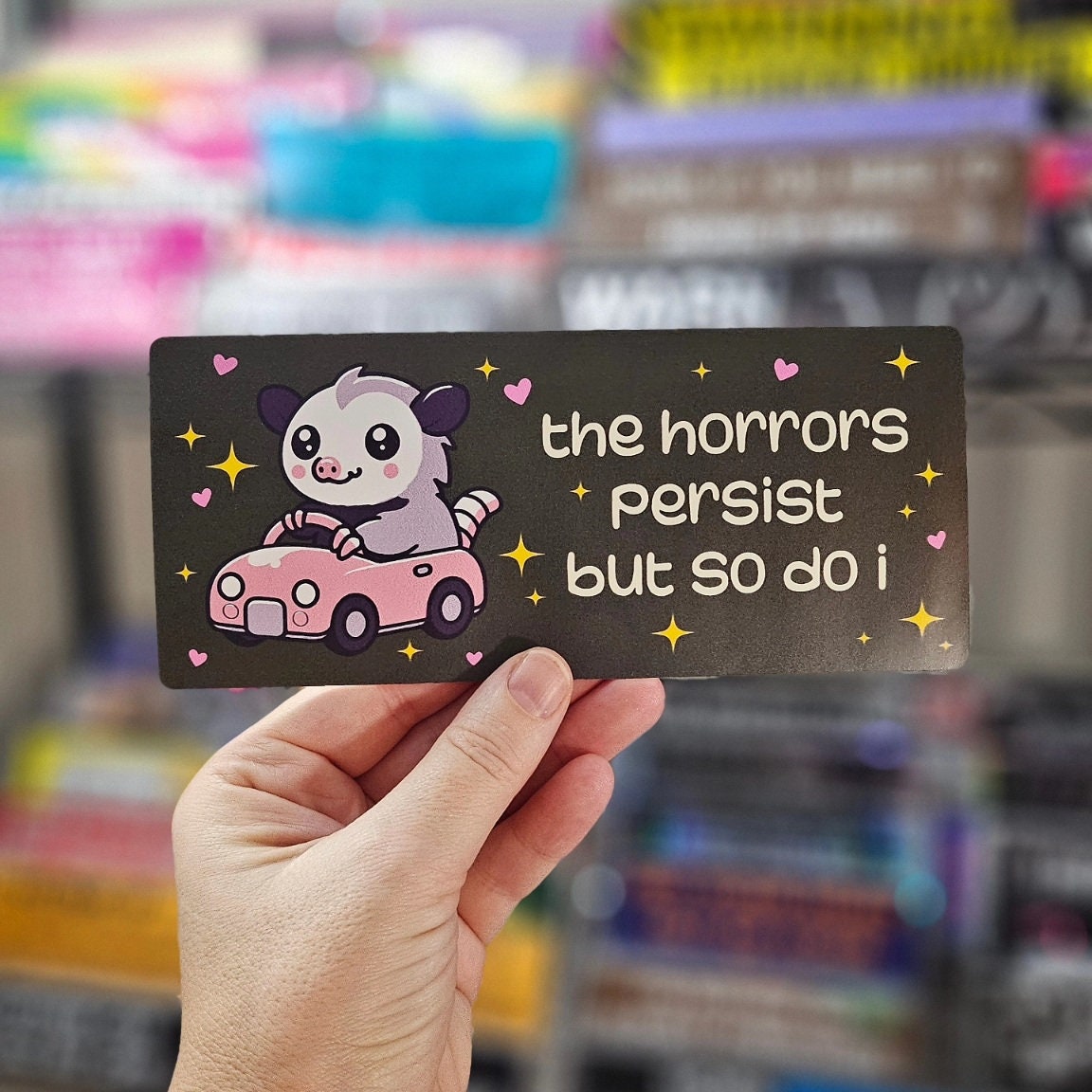 rectangular bumper stickers that say “the horrors persist but so do i,” featuring a cute possum driving a pink car with hearts and sparkles in pink, blue, and dark gray color options.
