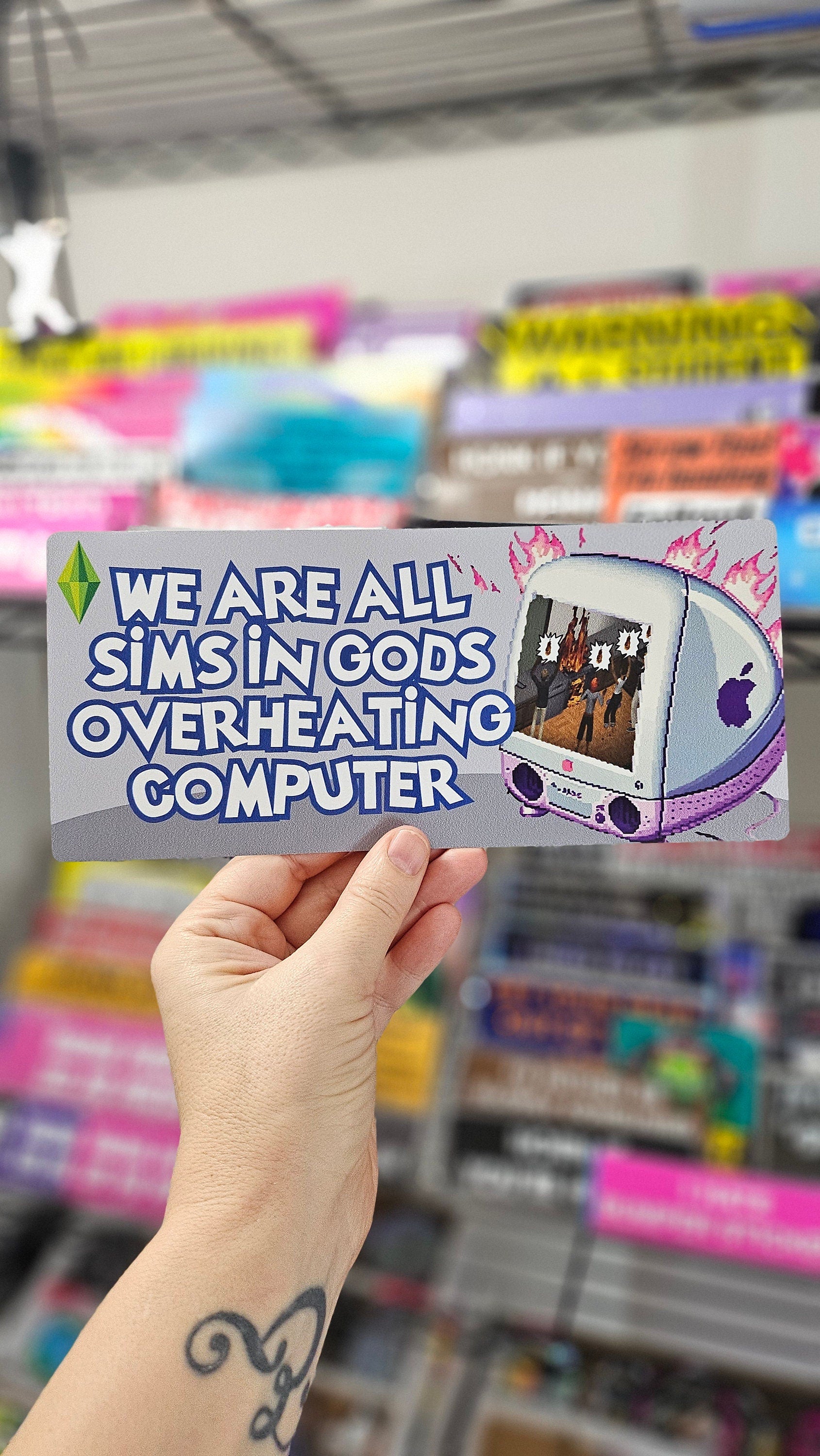 rectangular bumper stickers that say “We Are All Sims in God’s Overheating Computer,” featuring a retro computer, flames, and gamer-style artwork.

