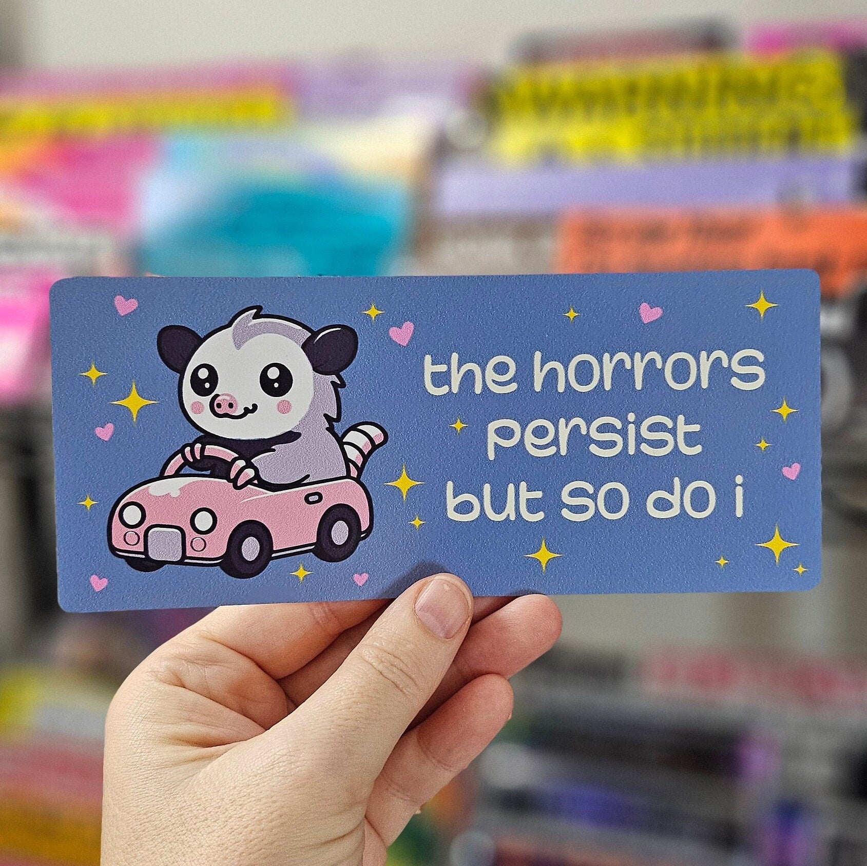 rectangular bumper stickers that say “the horrors persist but so do i,” featuring a cute possum driving a pink car with hearts and sparkles in pink, blue, and dark gray color options.
