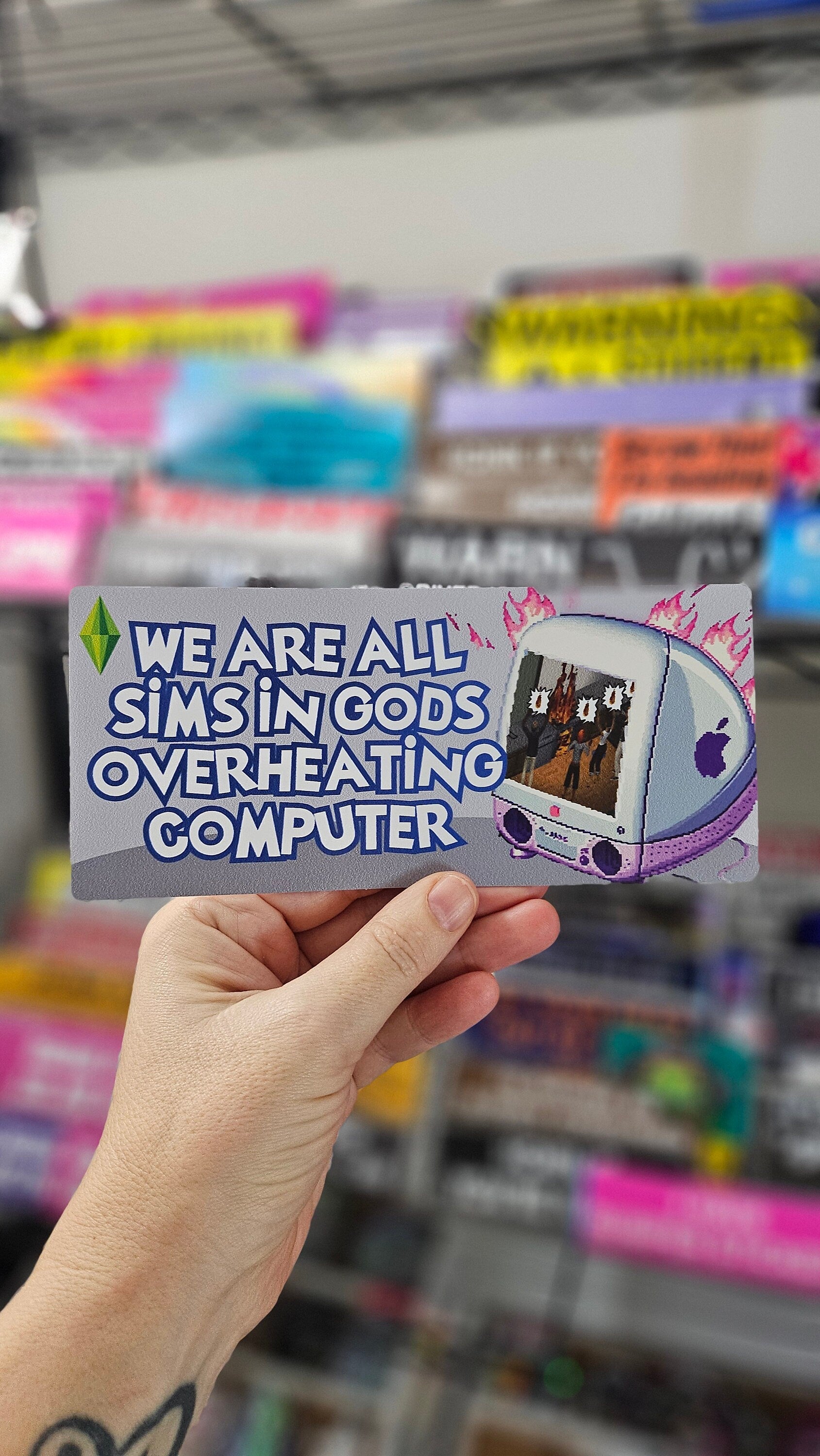 rectangular bumper stickers that say “We Are All Sims in God’s Overheating Computer,” featuring a retro computer, flames, and gamer-style artwork.
