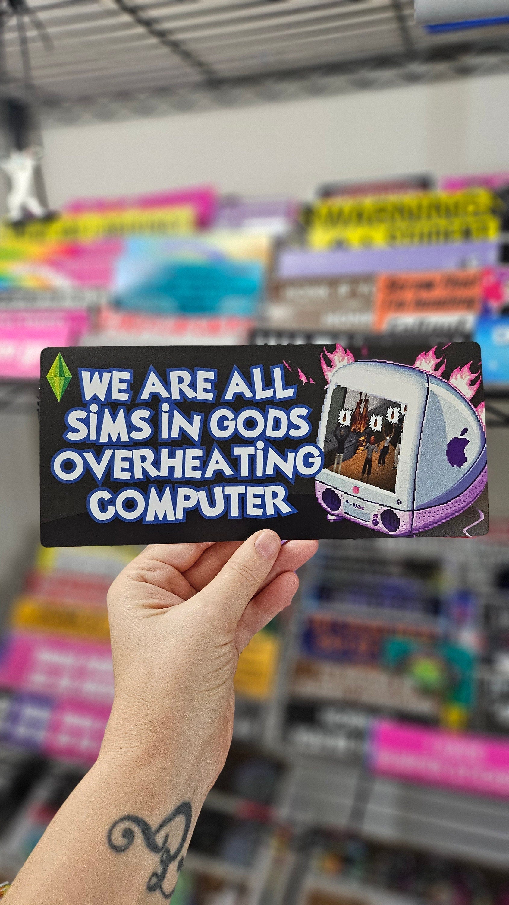 rectangular bumper stickers that say “We Are All Sims in God’s Overheating Computer,” featuring a retro computer, flames, and gamer-style artwork.
