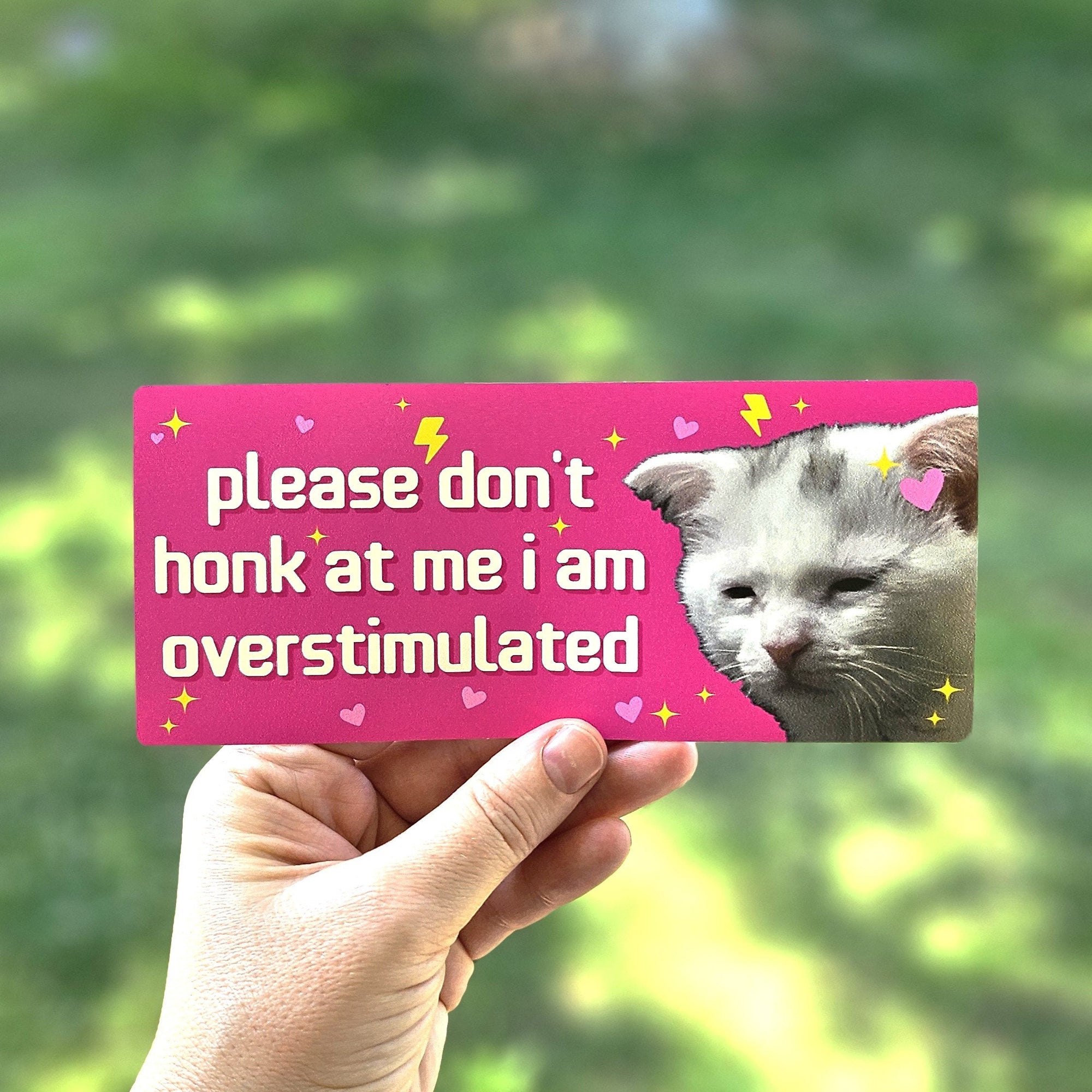 rectangular bumper stickers that say “please don’t honk at me i am overstimulated,” shown in pink and dark gray color options with a tired white cat, hearts, sparkles, and lightning bolts.
