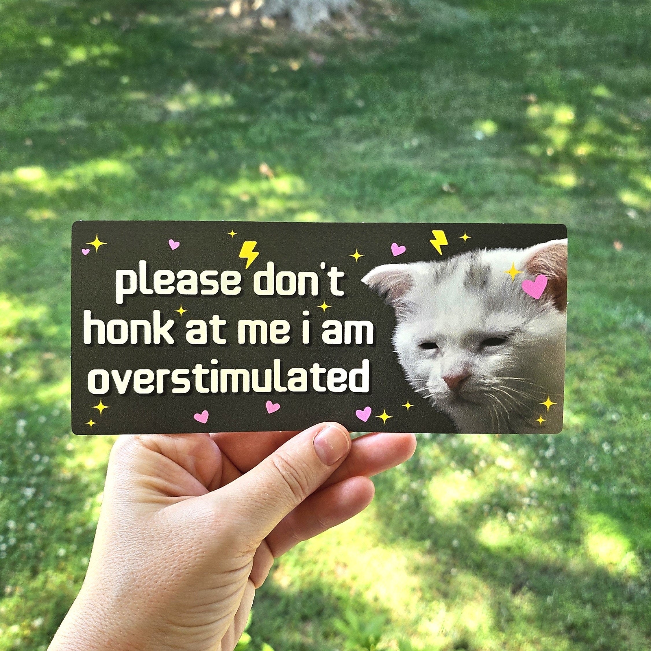 rectangular bumper stickers that say “please don’t honk at me i am overstimulated,” shown in pink and dark gray color options with a tired white cat, hearts, sparkles, and lightning bolts.
