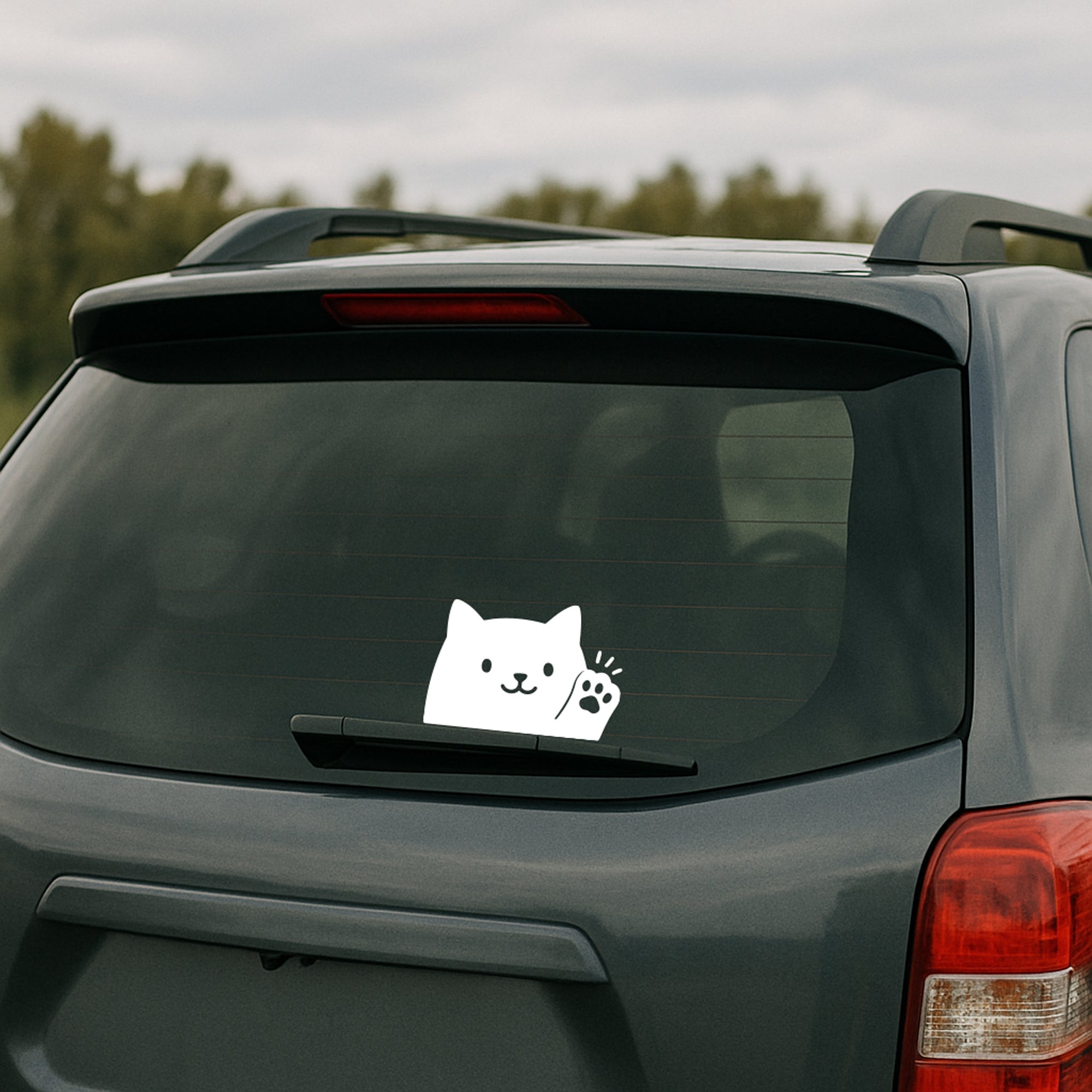 A white transfer decal of a cute cat waving hello or goodbye, applied on a car window with rain droplets.